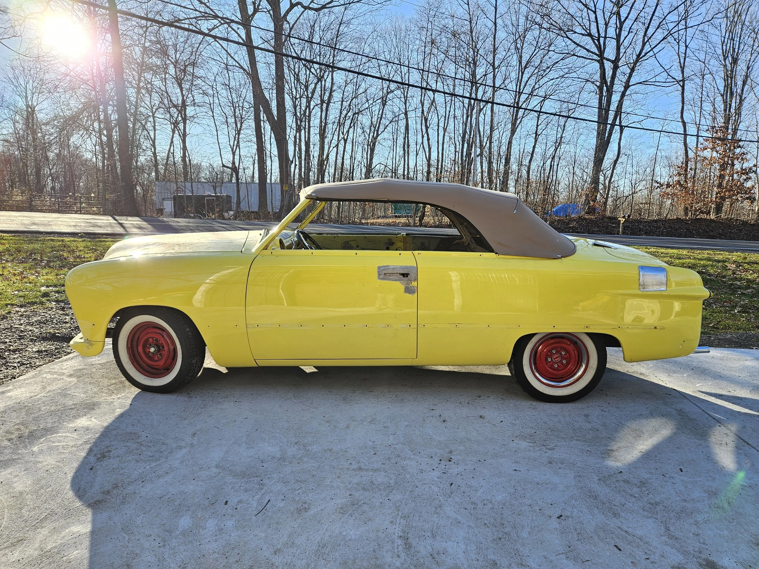 A yellow vintage convertible car with a beige soft top, parked on a concrete surface outdoors during daytime, with a background of leafless trees, power lines, and a clear blue sky.