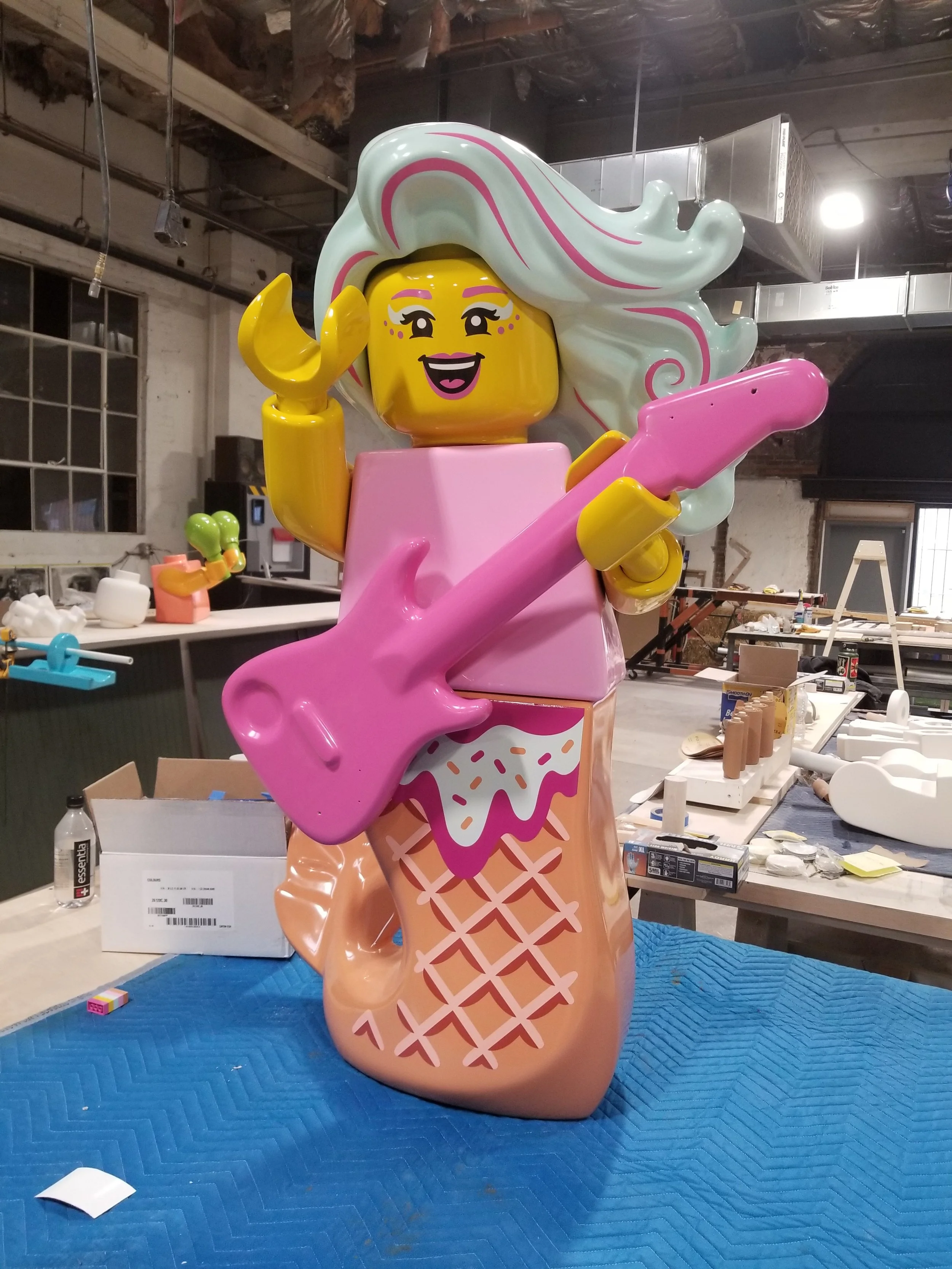 Colorful LEGO sculpture of a woman with long, wavy hair holding a pink guitar, wearing a pink top and ice cream cone skirt, in a workshop setting.