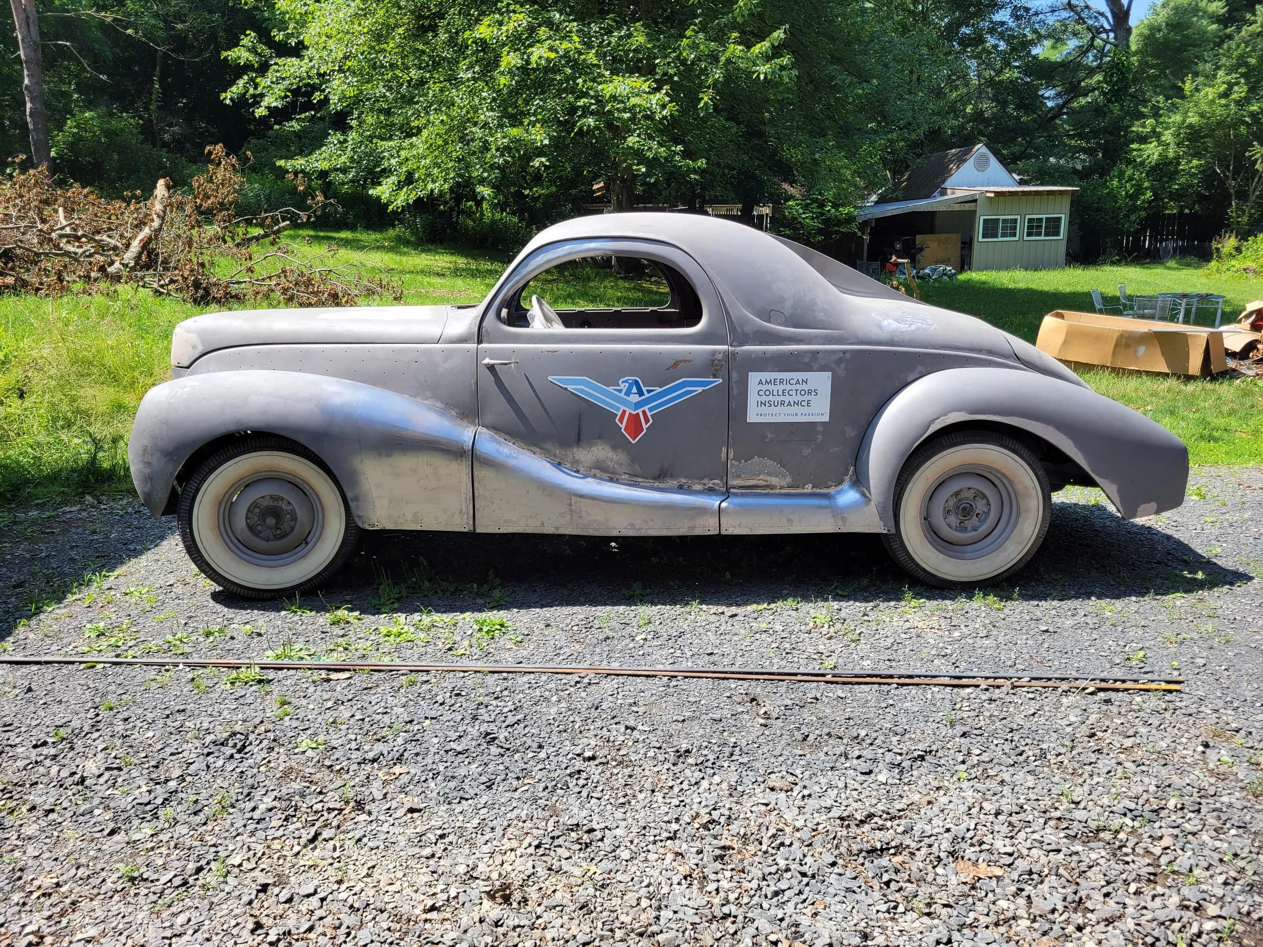 A vintage gray car with a Chevrolet logo and an American Collectors Insurance sign on the side, parked outdoors on gravel, with trees and a small building in the background.