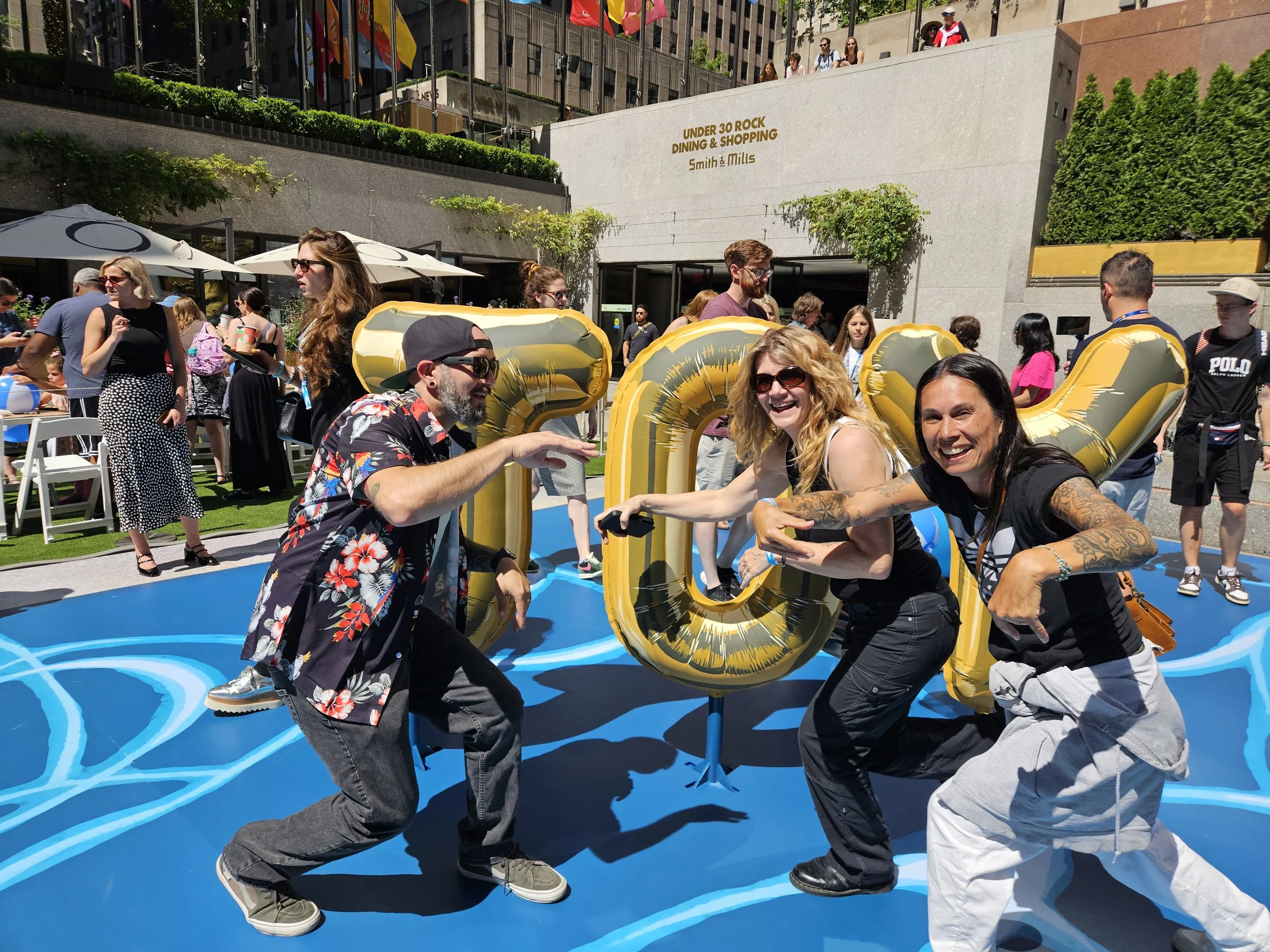 Four friends are posing playfully in front of large gold balloons spelling 'LOL' at an outdoor event on a sunny day, with many people in the background.