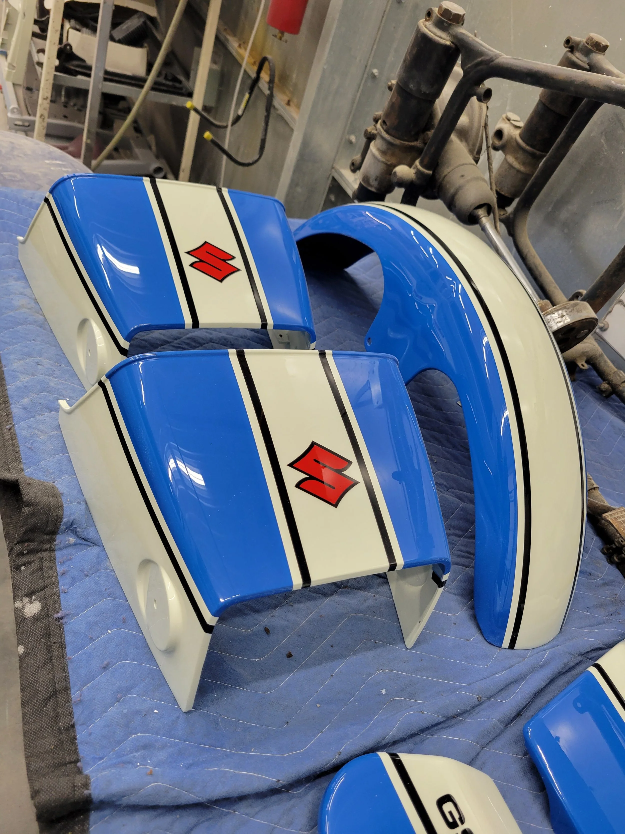 Blue and white motorcycle fairing with Suzuki logo, placed on a blue quilted surface in a workshop.