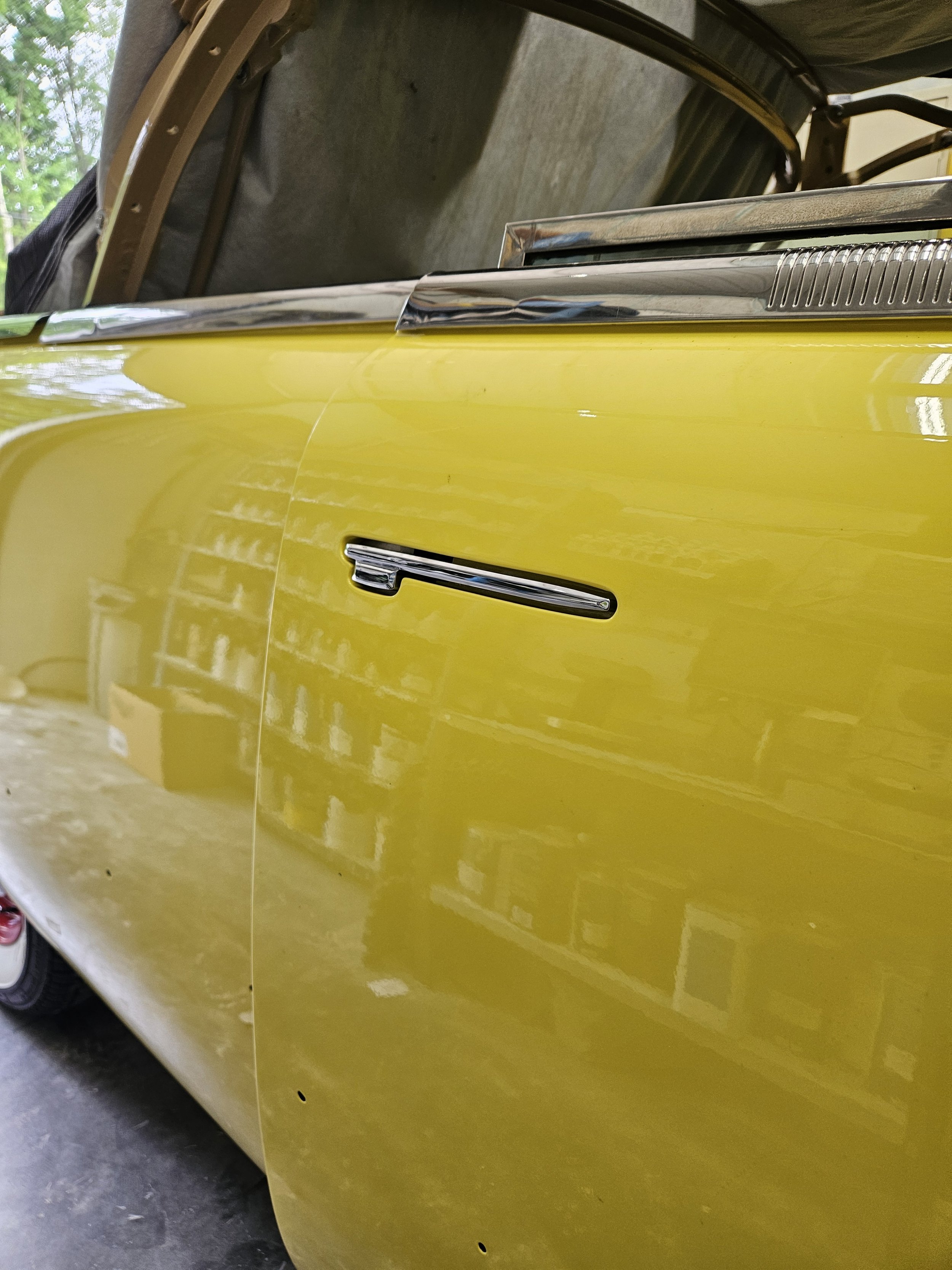 Close-up of a vintage yellow car door with chrome trim and a silver handle, reflected in the car's shiny surface.