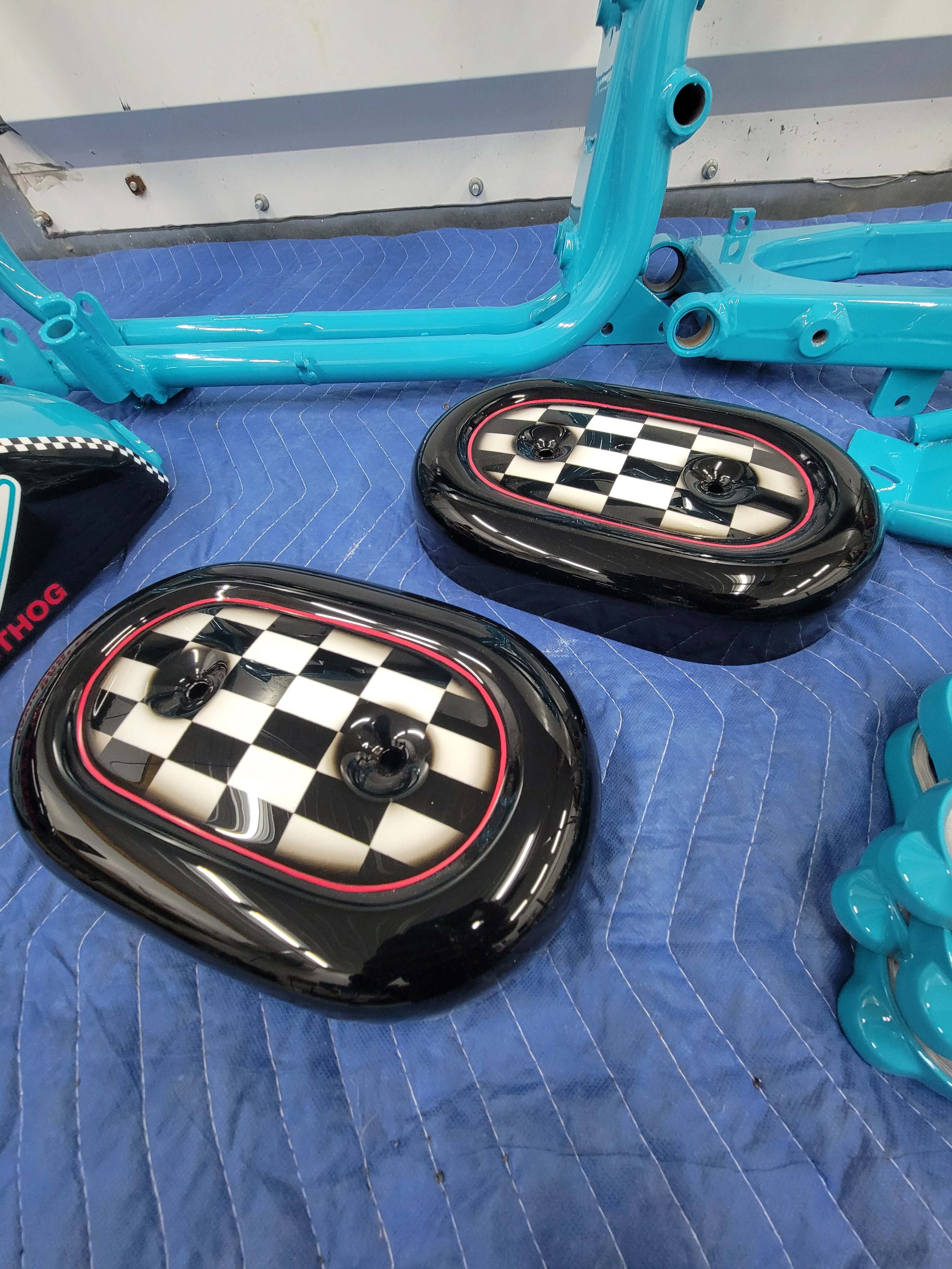 Disassembled parts of a bicycle, including a blue frame and black oval fenders with a white and black checkered pattern, on a blue padded work surface.