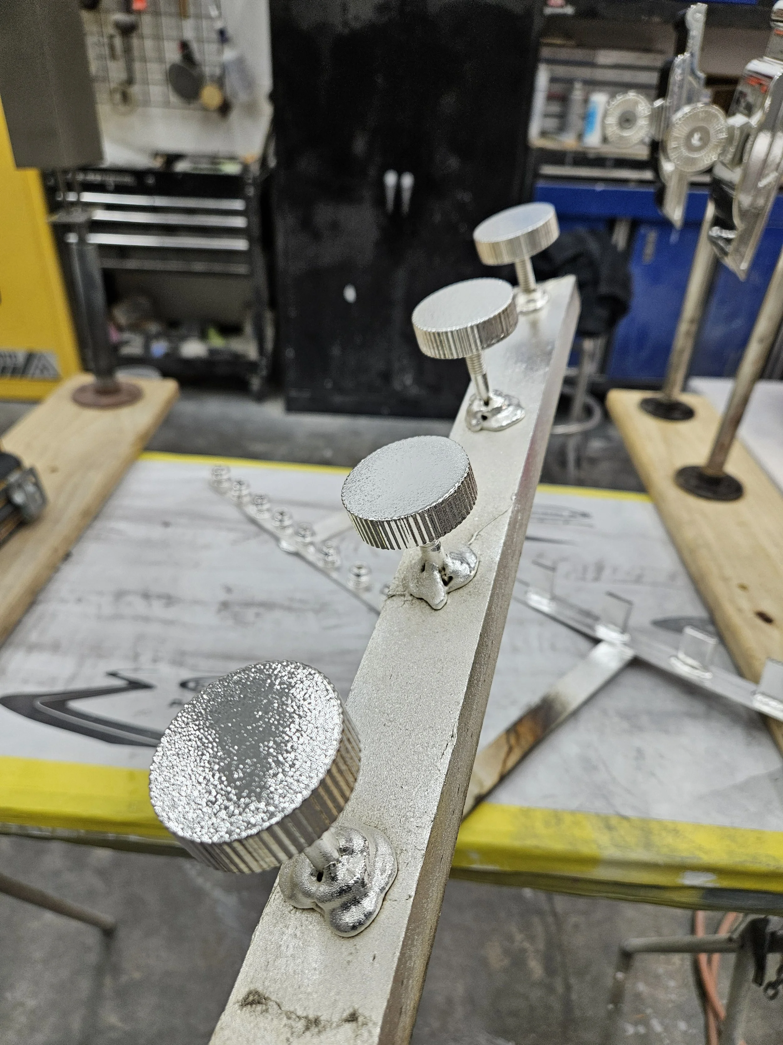 A metal workpiece with four knurled knobs fastened to it, mounted on a workbench in a workshop setting.