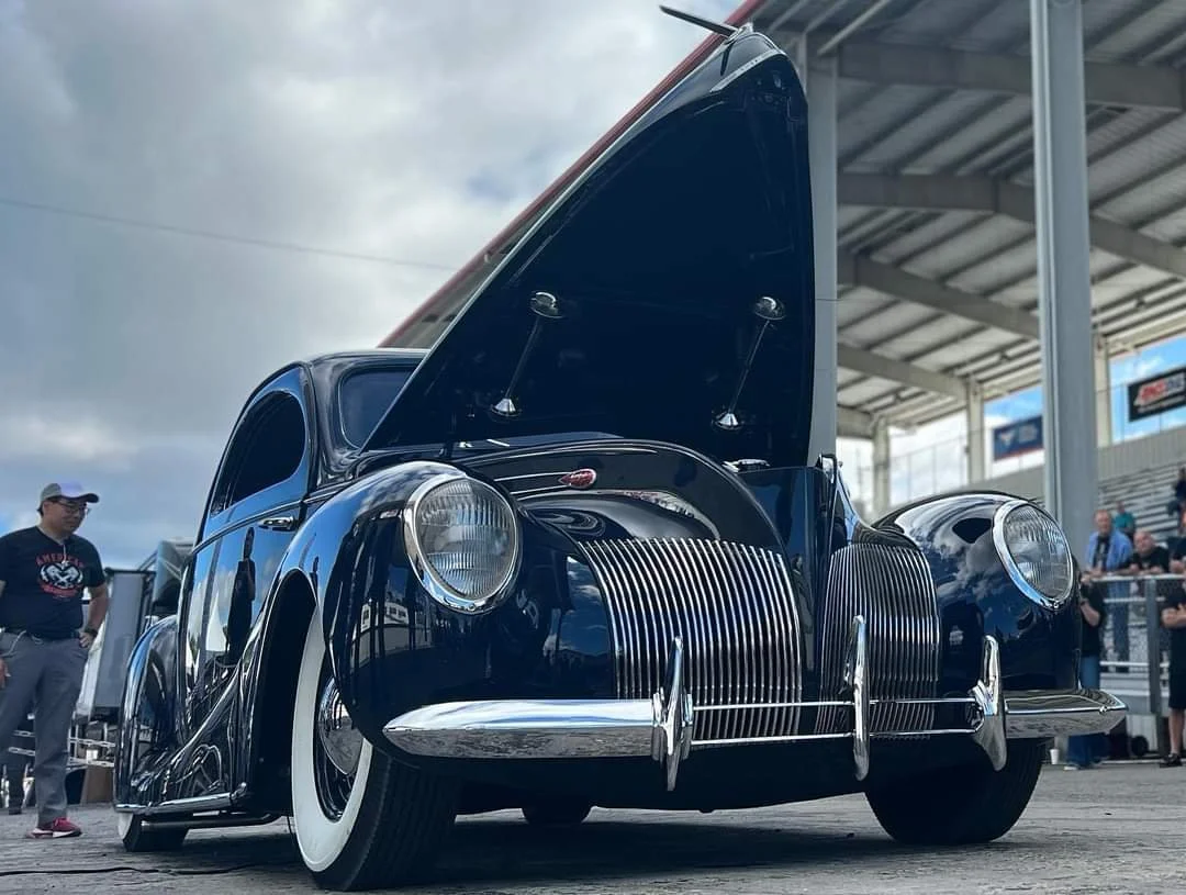 A vintage black car with its hood open at a car show, surrounded by people and a metal canopy structure.