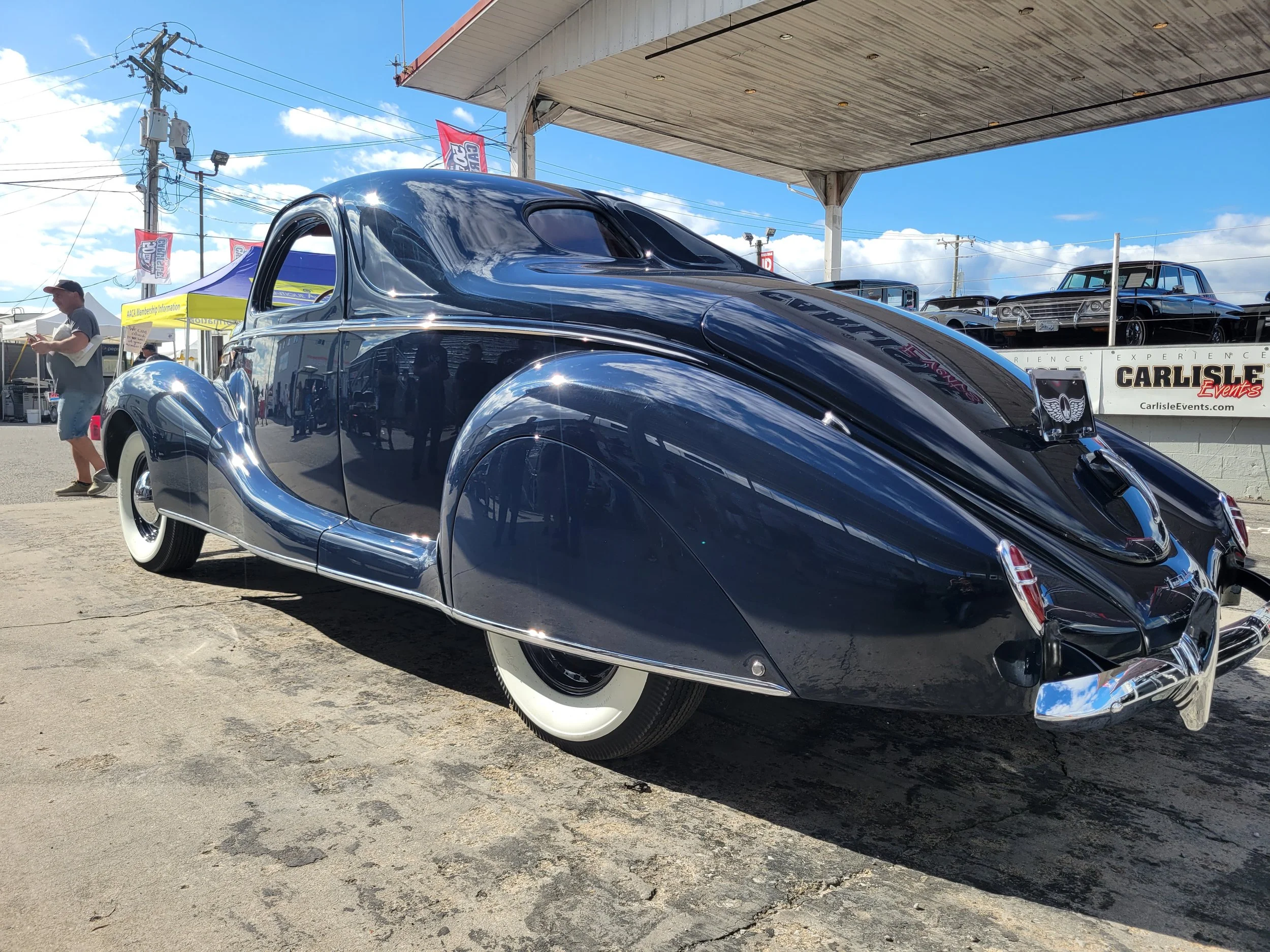 A shiny vintage black car with curved body and white wall tires parked at an outdoor car show.