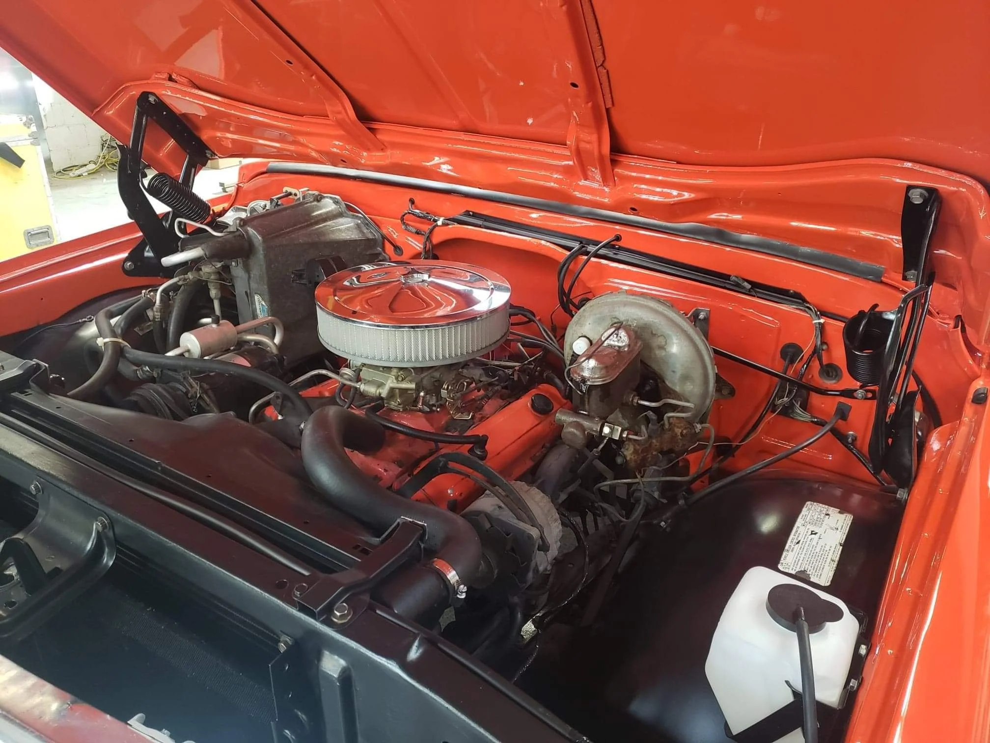 View of the engine bay of a vintage orange car, showing the engine with a chrome air cleaner, radiator, and various hoses and components.
