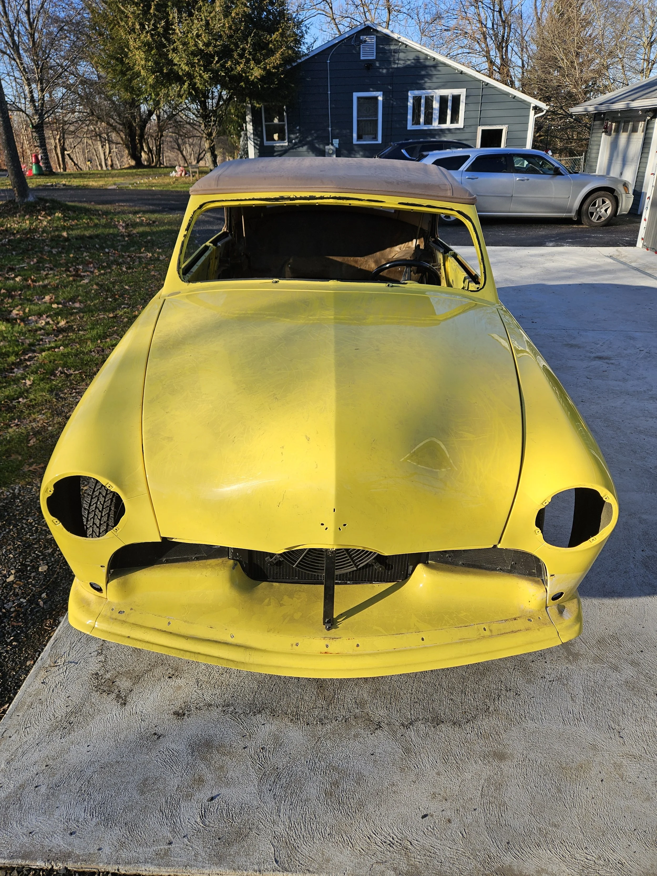 A yellow vintage car body without wheels or headlights, parked on a concrete area, with a house and two cars in the background.
