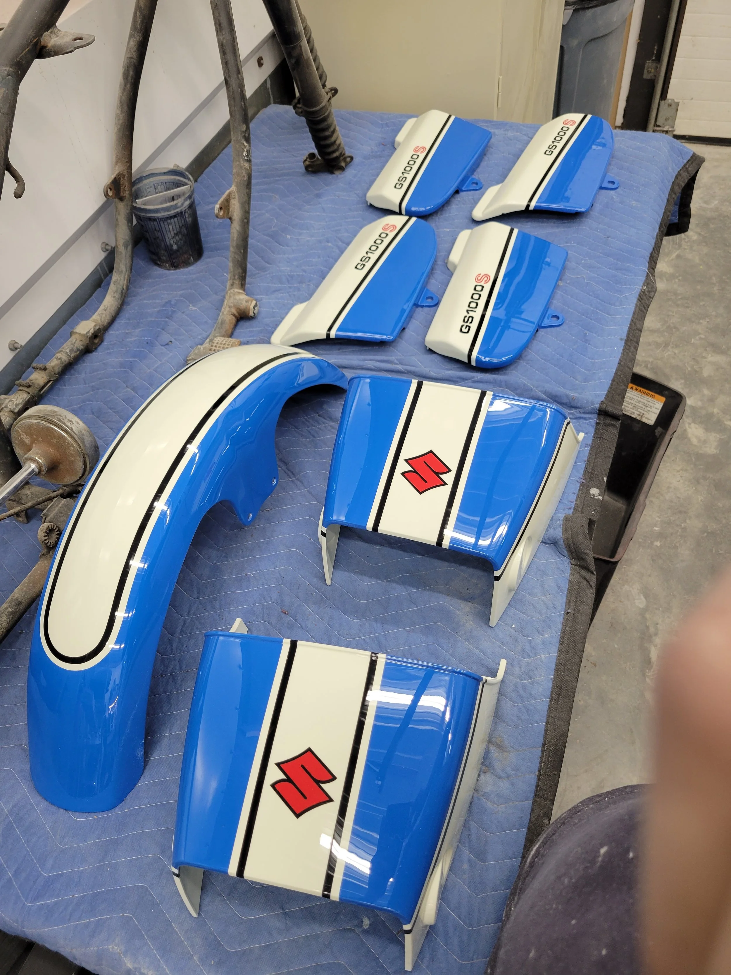 Collection of blue and white motorcycle fairing parts with Suzuki branding and red logo, arranged on a blue quilted surface in a workshop.
