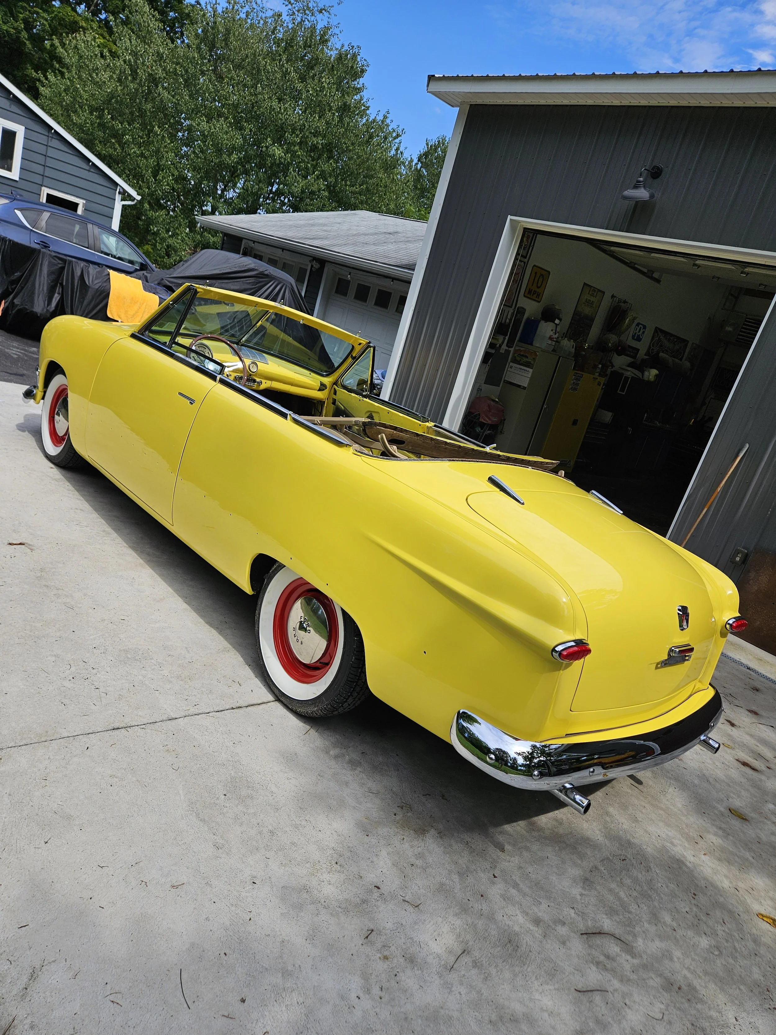 Yellow vintage convertible car parked outside a garage with an open door, revealing a workspace inside. The scene is set on a concrete driveway, with a covered car and some trees in the background.
