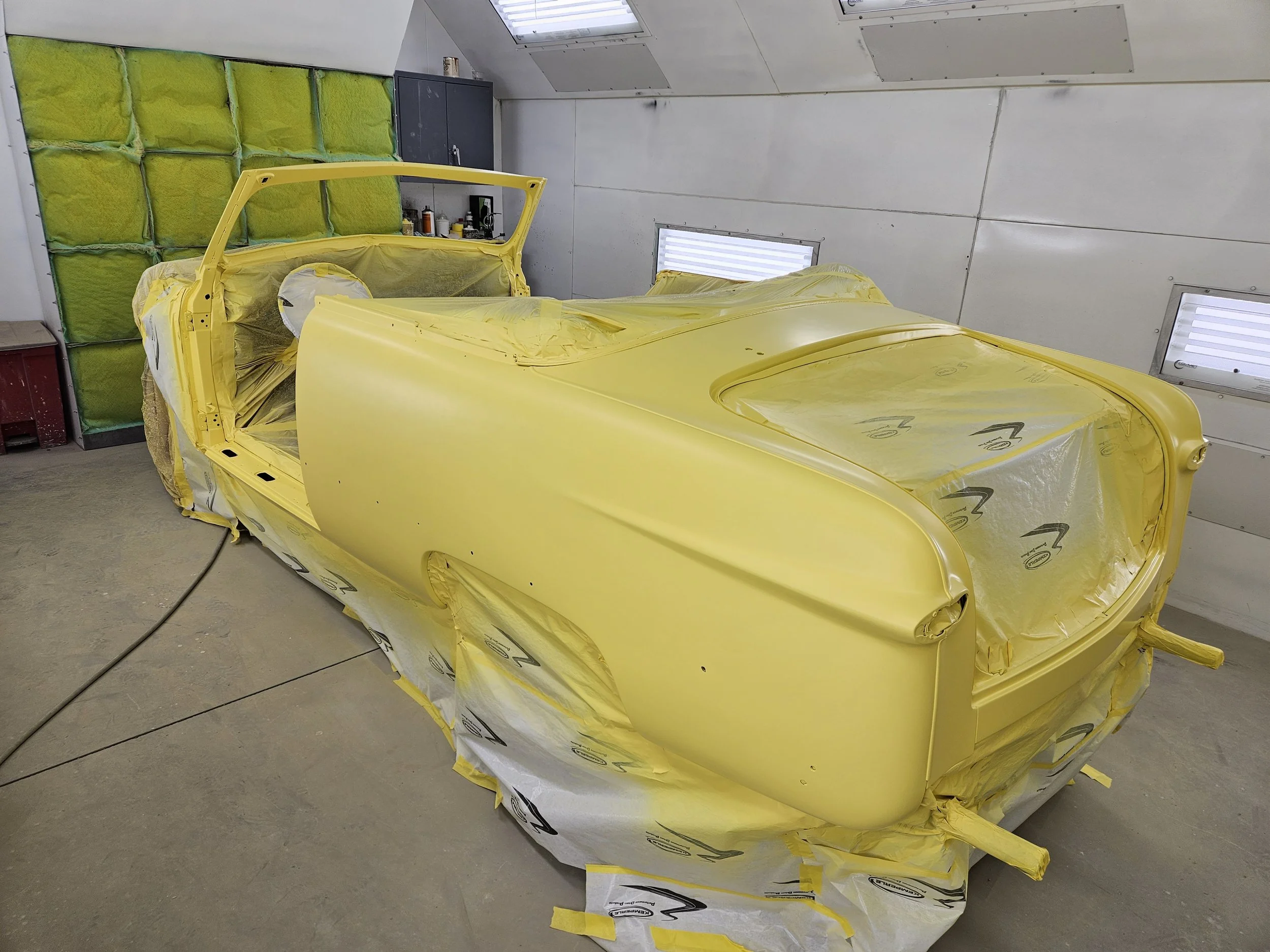 A yellow classic car is being painted inside a workshop, covered with masking paper and tape, with some paint supplies on a cabinet in the background.