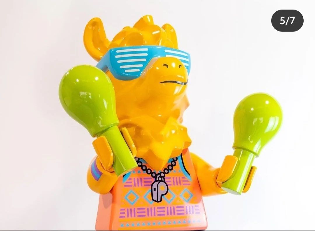 Plastic LEGO sculpture of a yellow camel wearing blue sunglasses, a colorful shirt, and green boxing gloves, set against a plain white background.
