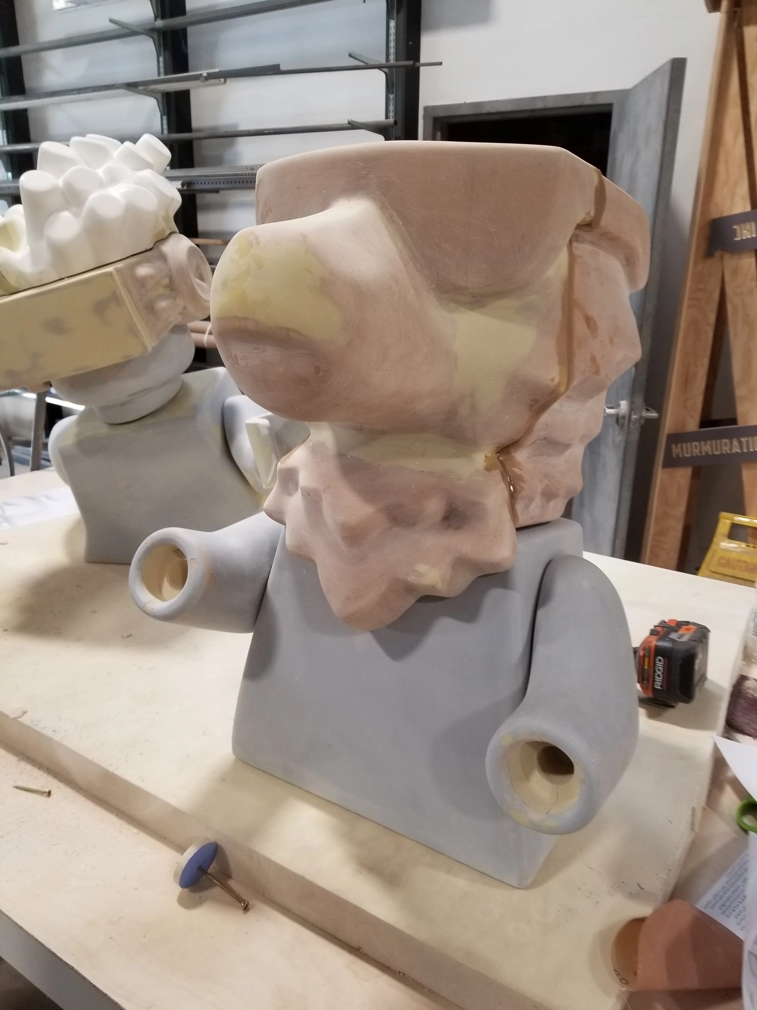 LEGO sculpture made of unfinished plaster or ceramic, sitting on a gray base in a workshop surrounded by tools and other sculptures.