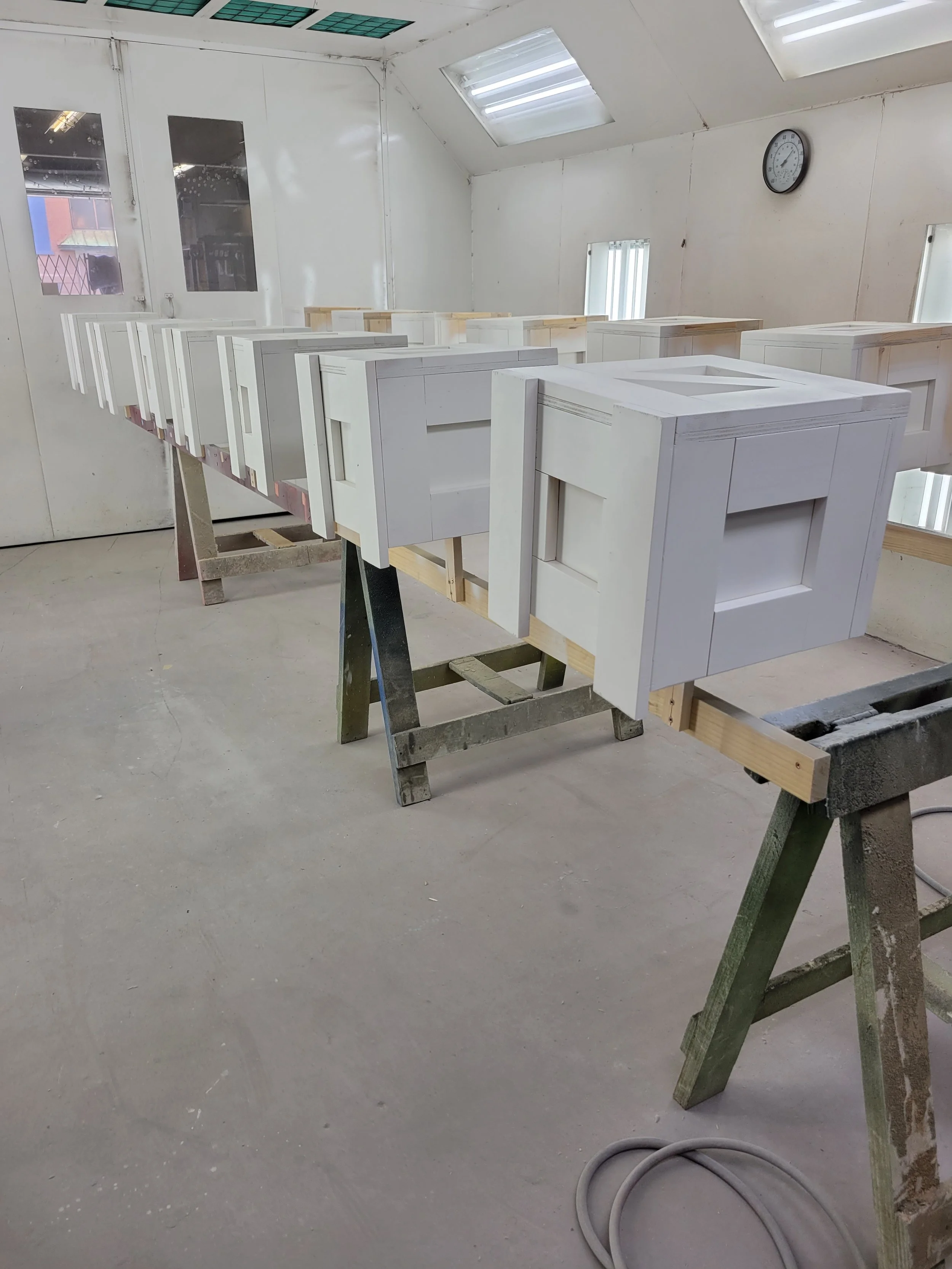 Multiple white cabinet frames in a woodworking shop on workbenches with a white wall, a wall clock, and skylights in the ceiling.