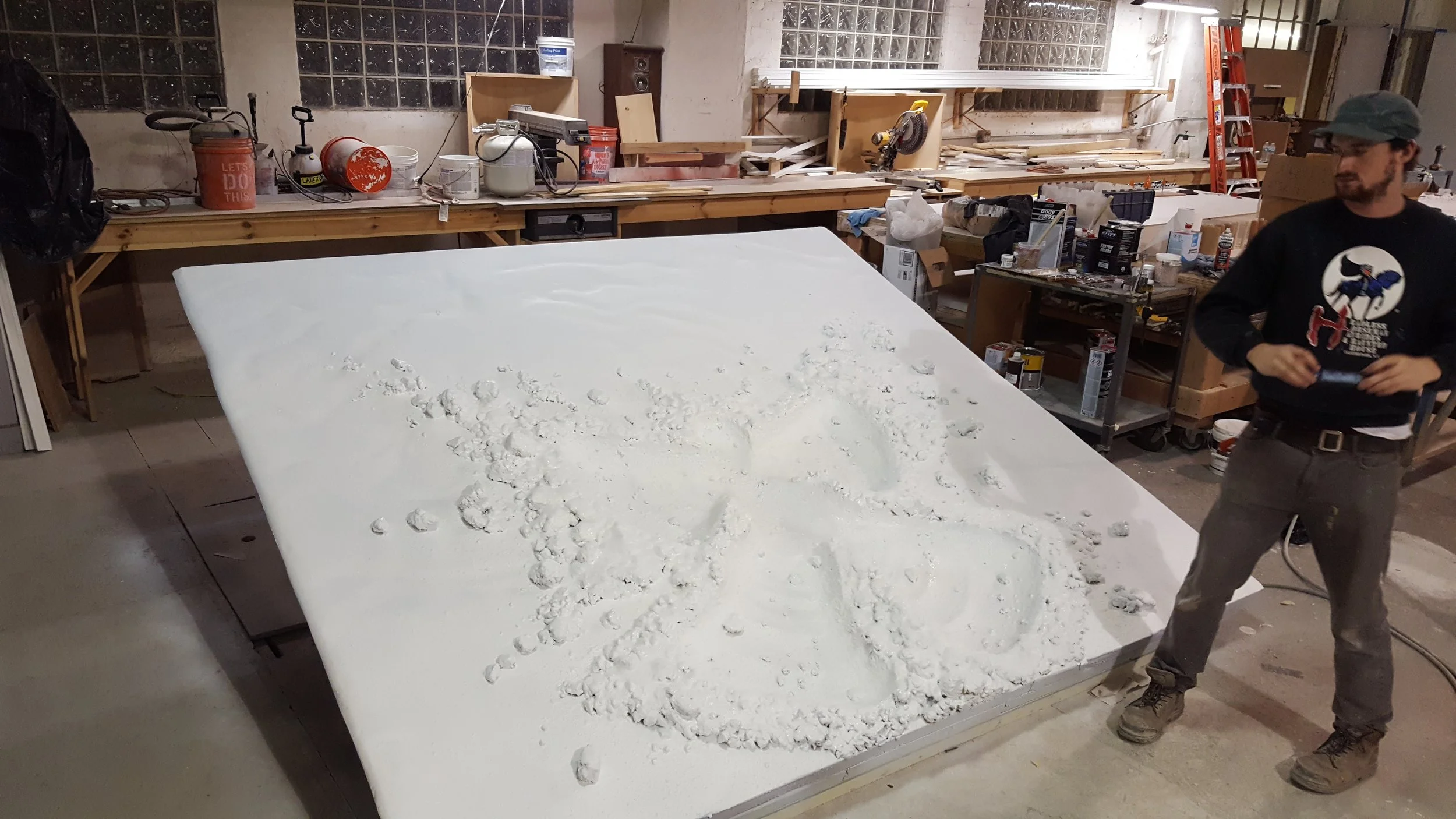 A workshop with a large white surface replicating a snow angel. The background has various tools, supplies, and a ladder.