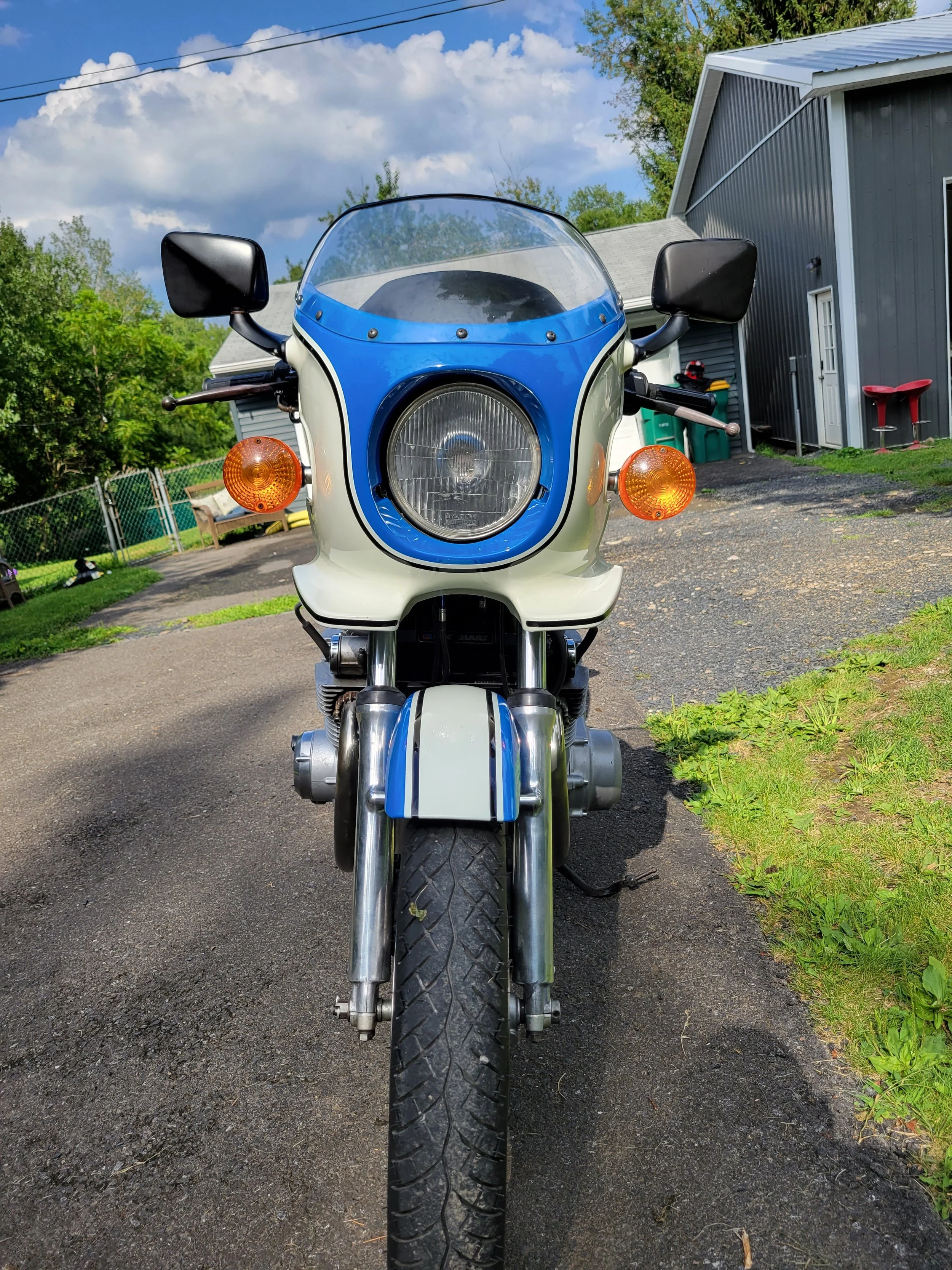 Front view of a vintage motorcycle with a white and blue color scheme, parked on a paved driveway in front of a gray building with trees and a bench in the background.