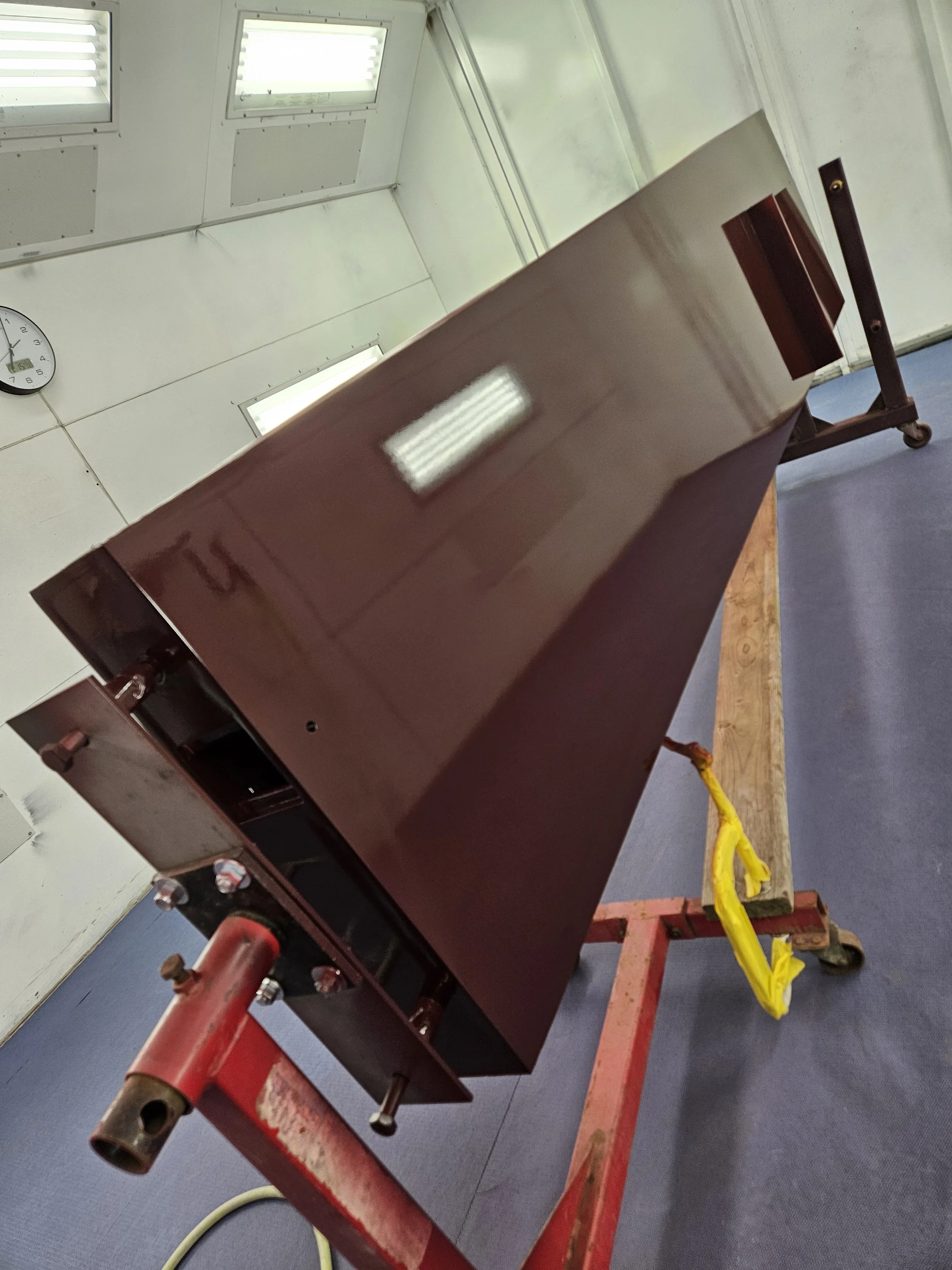 A large, rectangular, brown metal box on a red wheeled cart in a room with white walls and ceiling, fluorescent lights, and a clock showing 10:03.