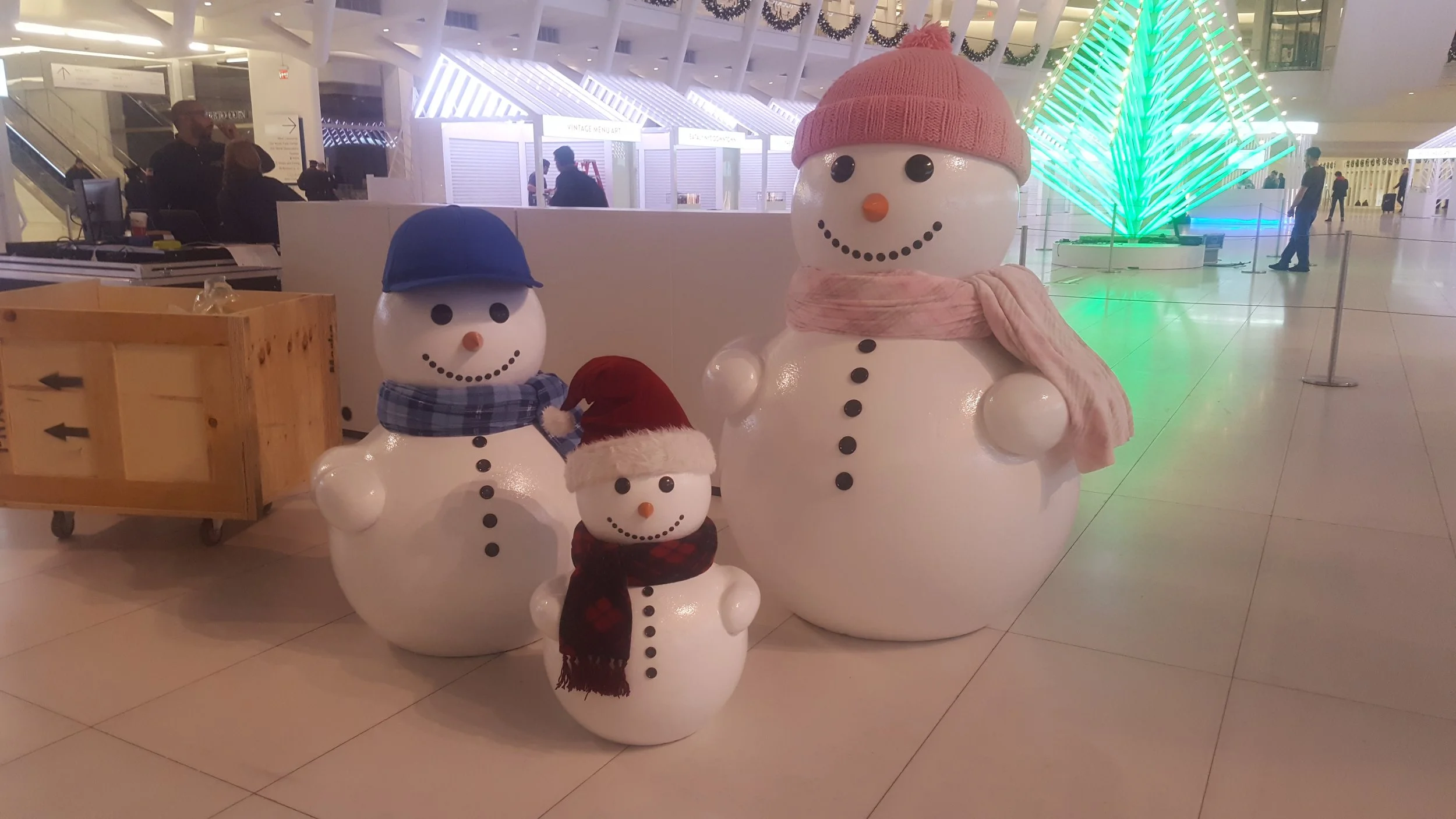 Three snowman figures decorated with winter accessories, inside a modern, well-lit building. The largest snowman wears a pink knit hat and scarf, the medium-sized snowman wears a blue cap and checkered scarf, and the smallest snowman, wearing a Santa