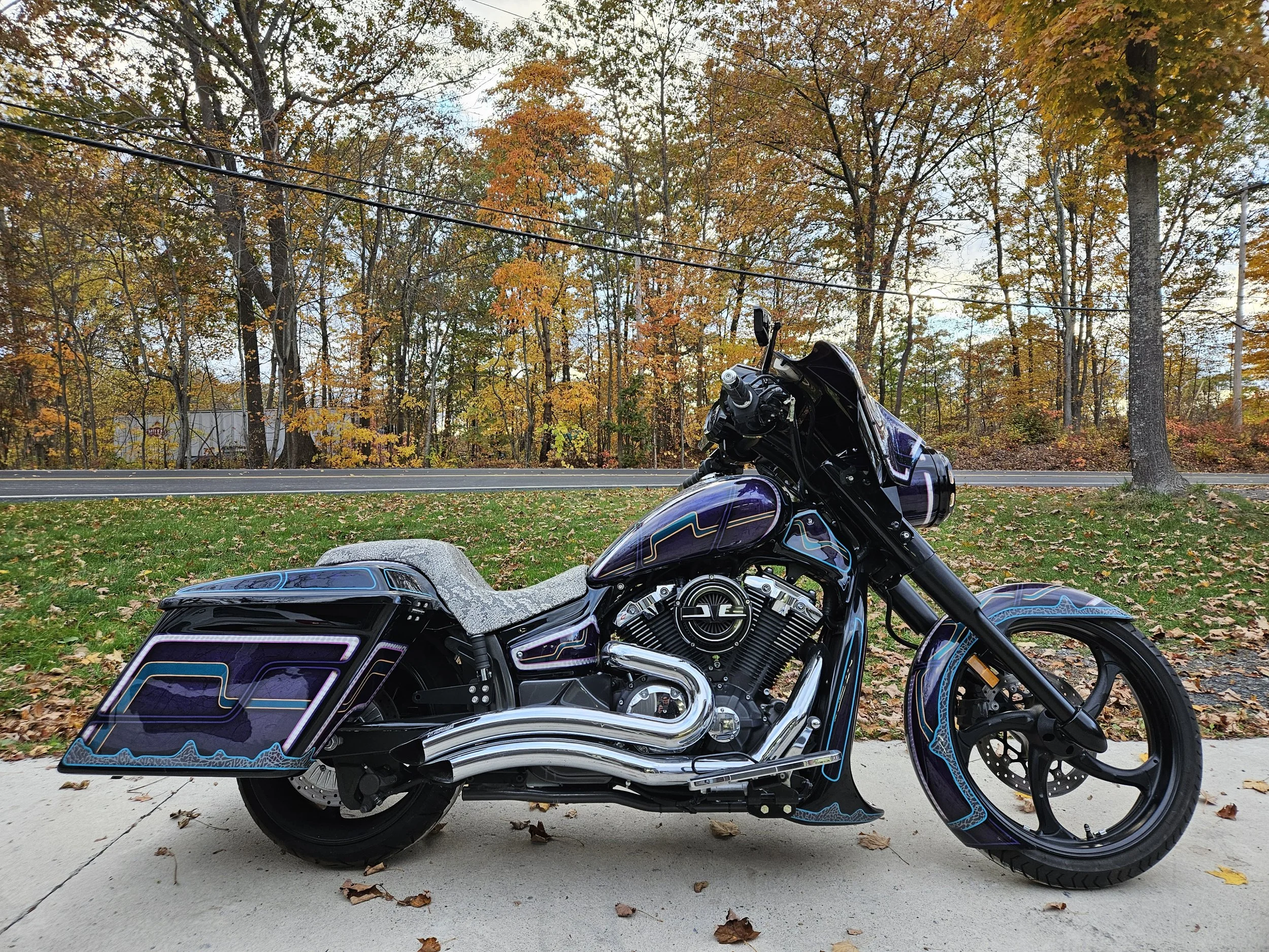 A custom motorcycle with intricate blue and purple graphics parked on a concrete driveway with autumn leaves scattered around, in front of a background of trees with fall foliage.
