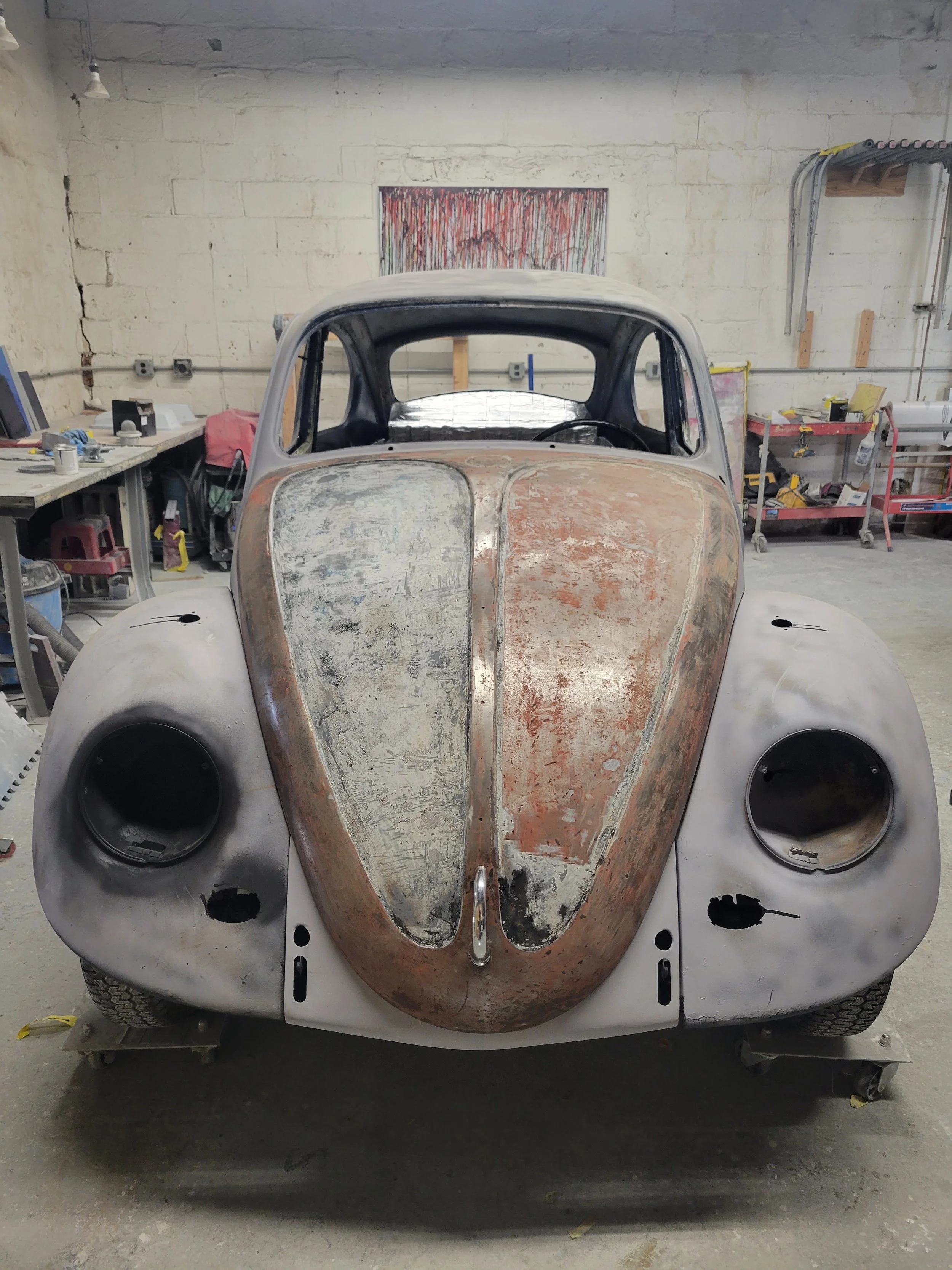 Front view of a vintage car chassis undergoing restoration in a workshop, with a partially sanded and rusted body, missing headlights and other parts.
