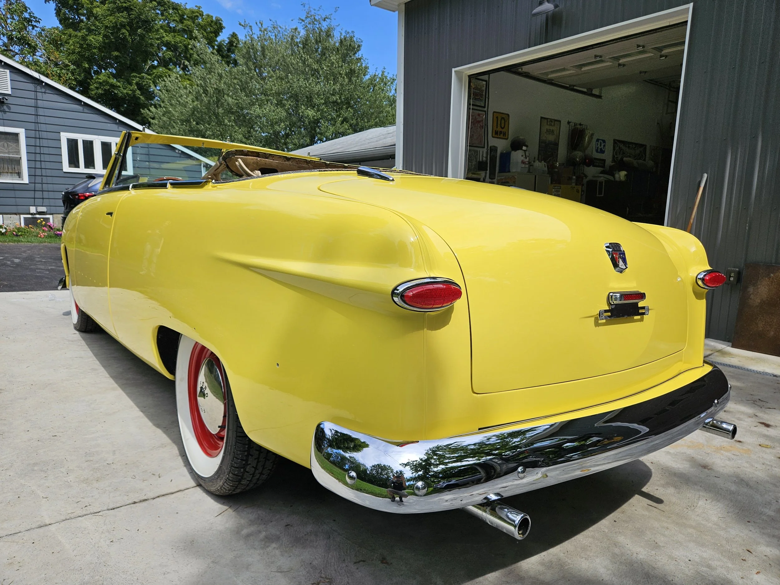 Yellow vintage convertible car parked outside a garage, with reflections on chrome bumpers and red details on wheels.
