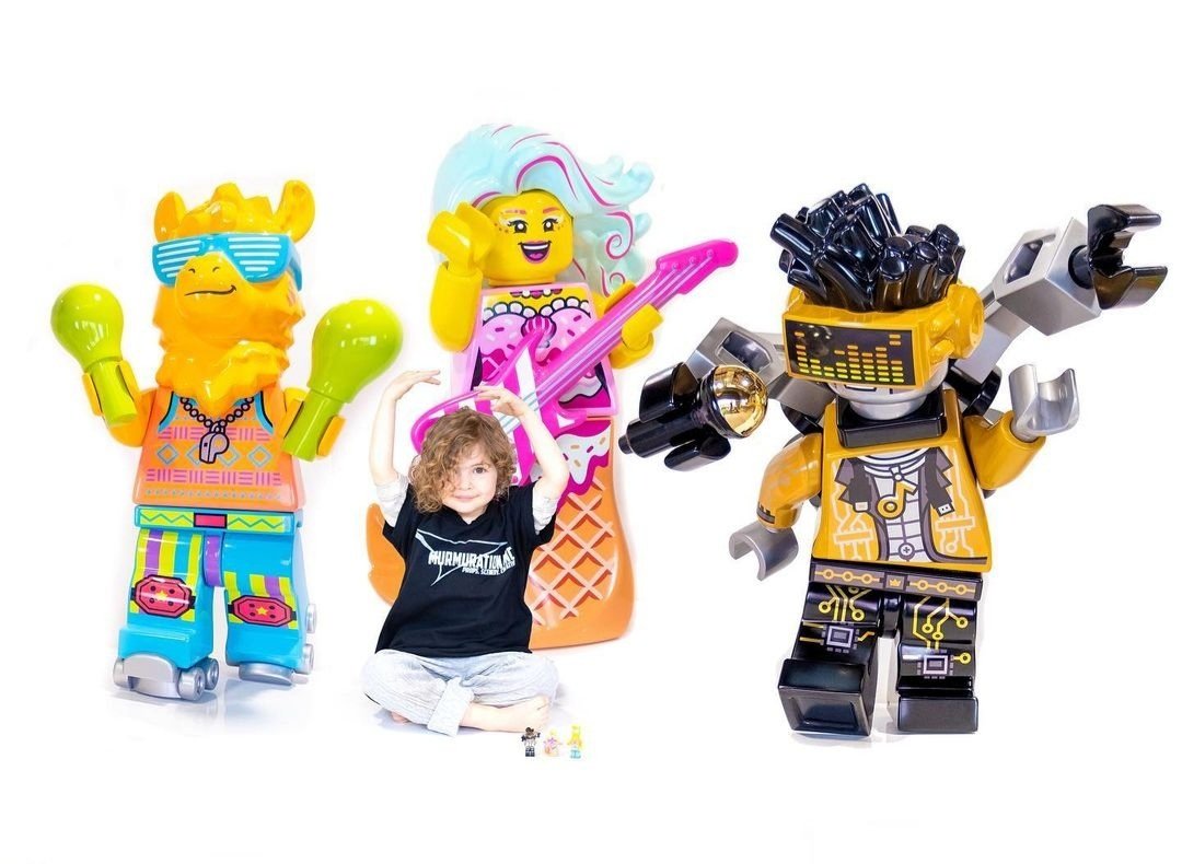 A child sitting on the floor with three colorful Lego characters: one with orange hair, sunglasses, and sports attire; another with long, flowing, multicolored hair, and a pink guitar; and a third with black and yellow armor, glasses, and a robotic a