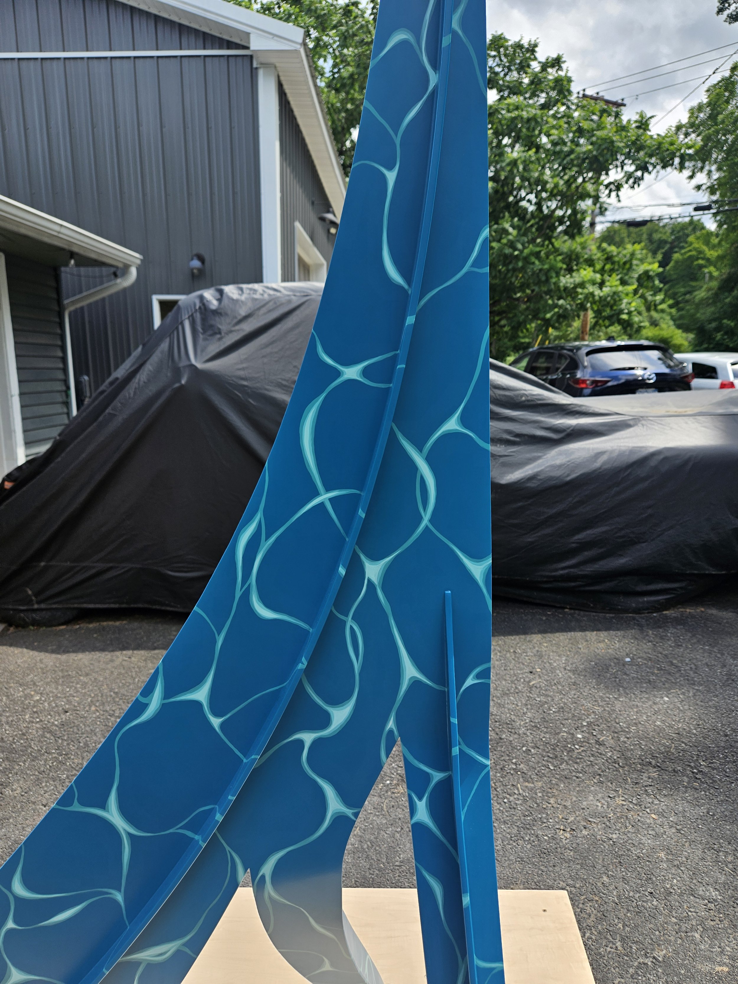 A blue sculpture with a water ripple pattern leaning against a wooden stand outdoors, with a black tarp-covered object, a house, and trees in the background.