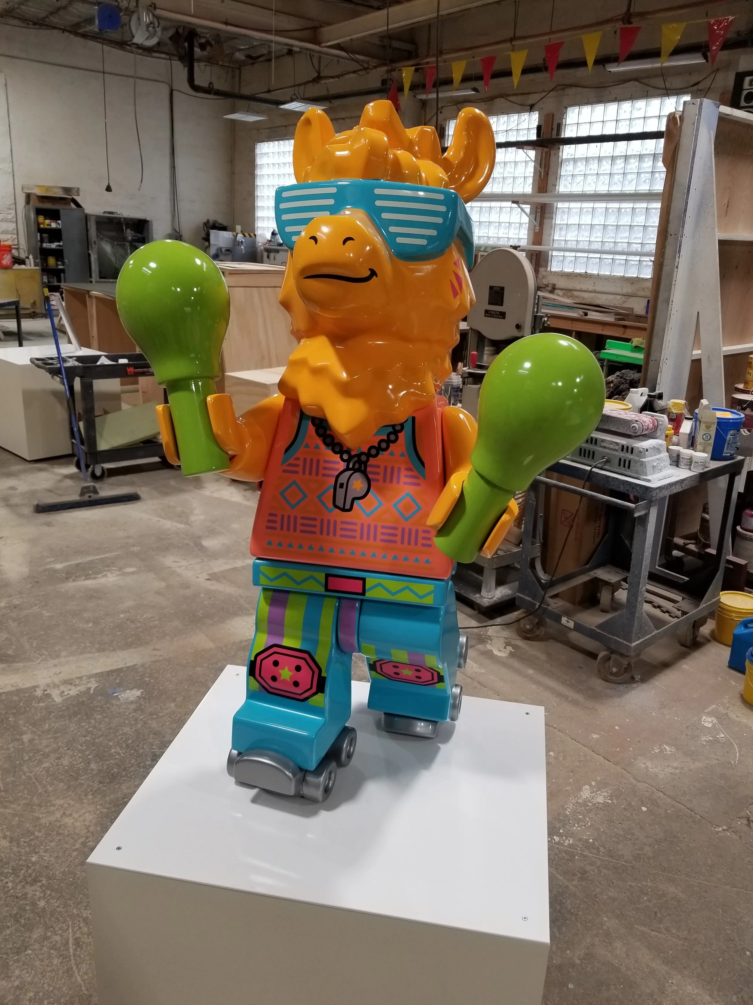 Colorful LEGO camel sculpture wearing sunglasses and a necklace, displayed indoors with workshop tools and furniture in the background.
