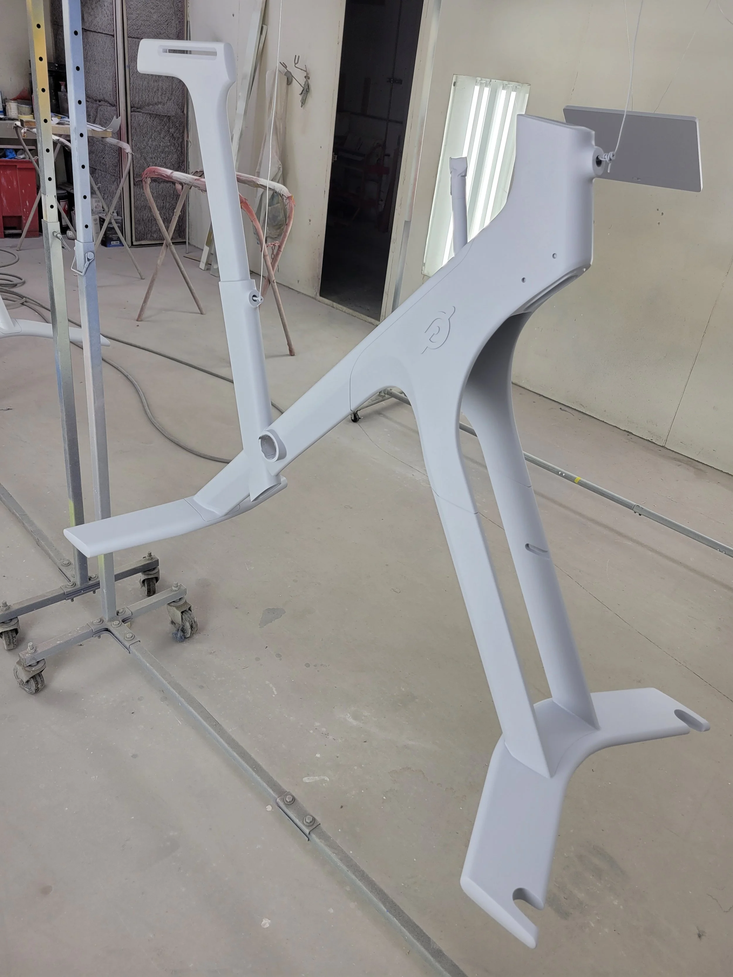 White bicycle frame in a workshop setting, mounted on a stand, with tools and equipment around.