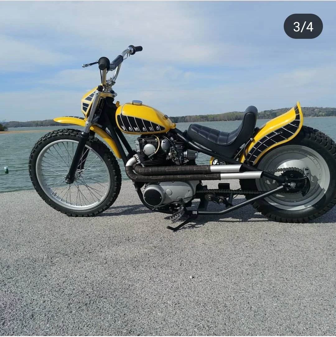 A custom yellow motorcycle with black accents and large tires is parked on a paved surface near a body of water, with hills and a cloudy sky in the background.
