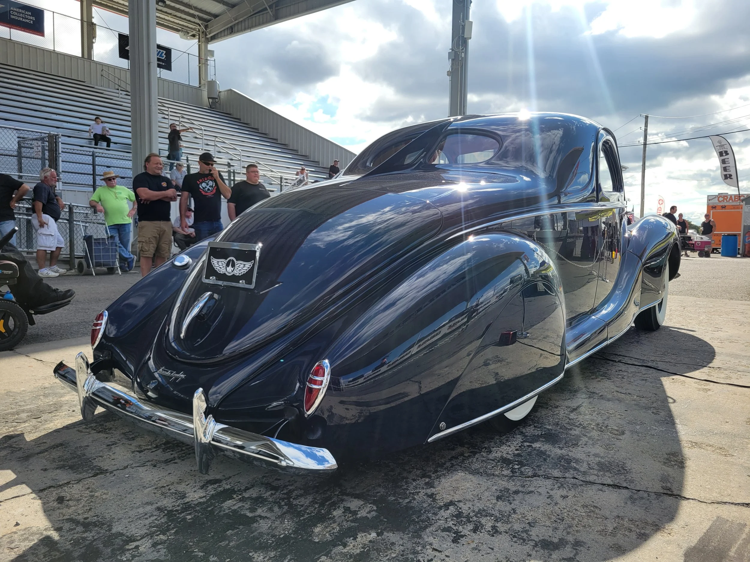 A vintage black car at an outdoor event with people standing and sitting on bleachers in the background, partly cloudy sky, and food stalls visible.
