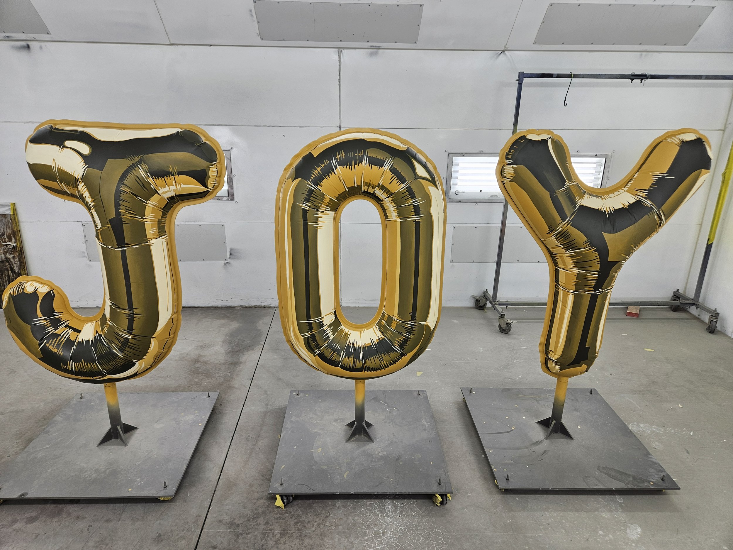 Large metallic balloons forming the word 'JOY' in black and gold, standing on a grey floor in a room with white walls and fluorescent lights.
