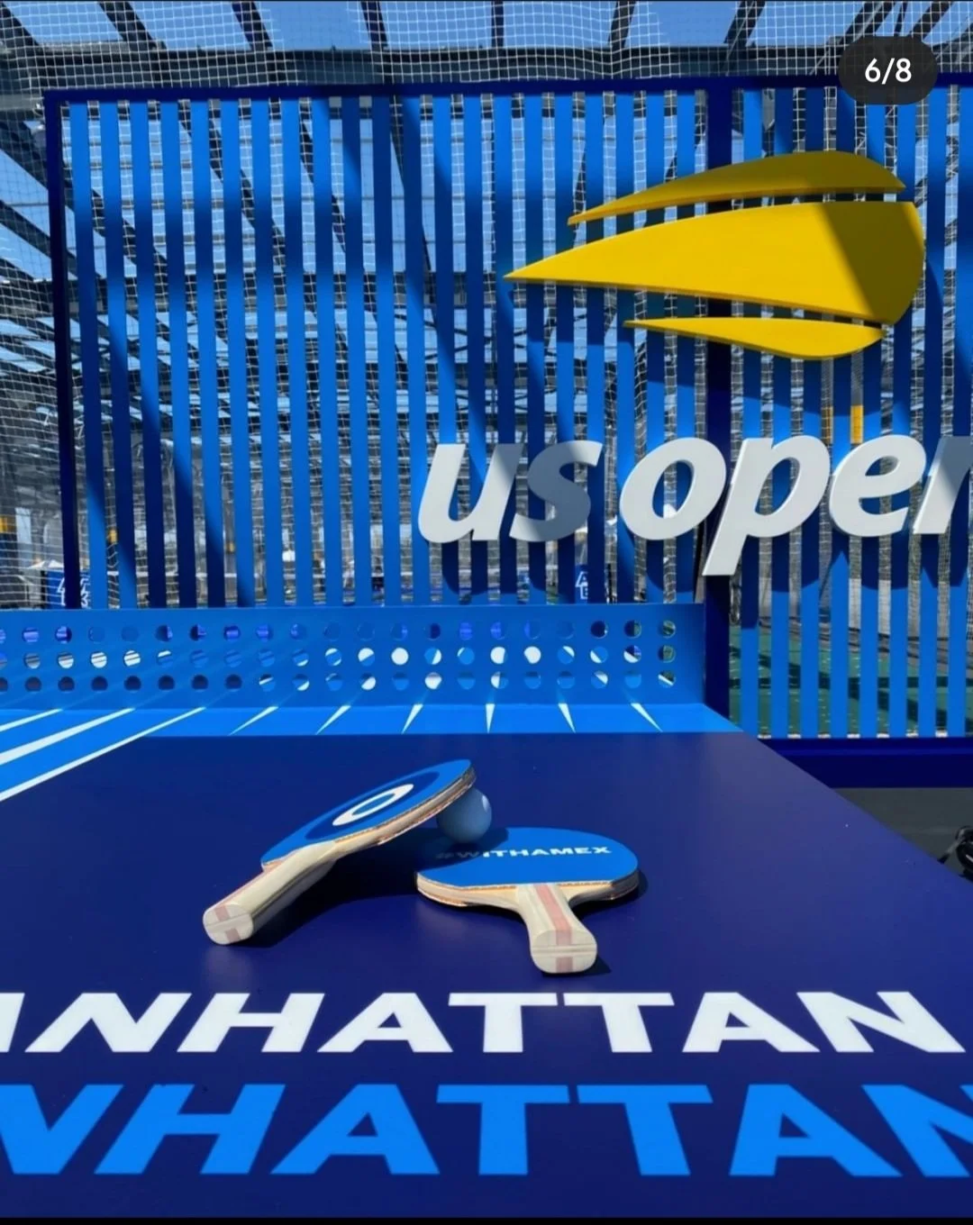 Ping pong paddles and ball on a table tennis table with 'usopen' and 'WHATTAN' branding, set up inside a fenced area with blue structural elements.