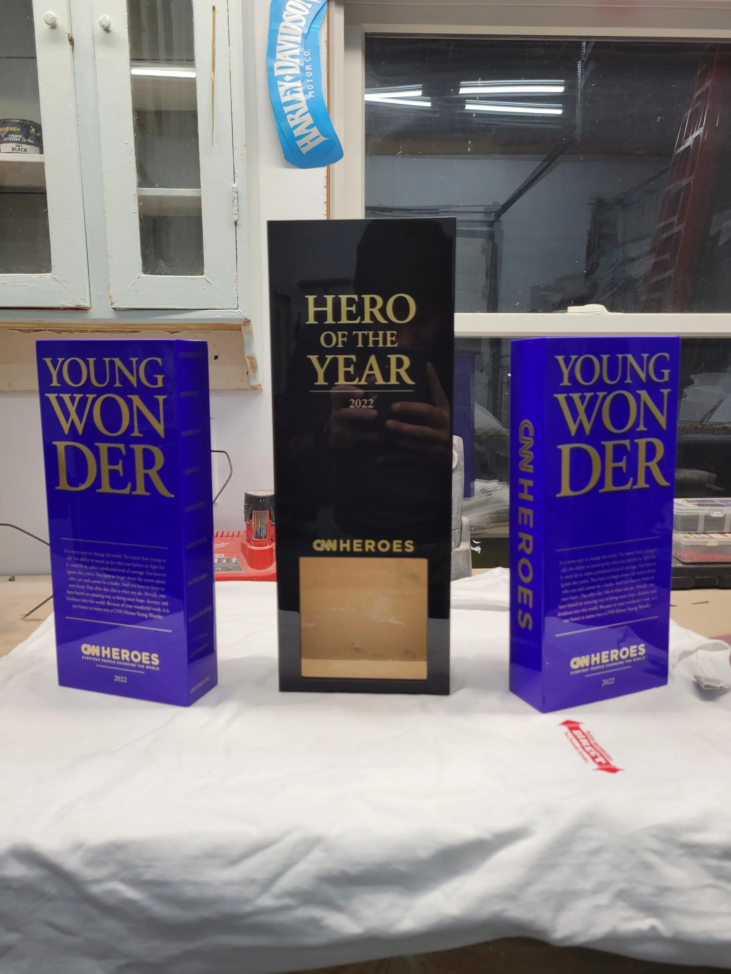 A black award trophy with gold lettering that reads 'Hero of the Year 2022 CNN Heroes' is displayed on a table alongside two blue boxes labeled 'Young Wonder'. The table is covered with a white cloth, and the background shows a workshop or garage wit