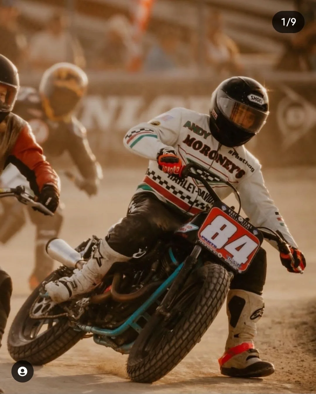 Motocross racers leaning into a turn on a dirt track during a race, the rider in foreground wearing a helmet and racing gear with Harley-Davidson branding and the number 12 on the bike.