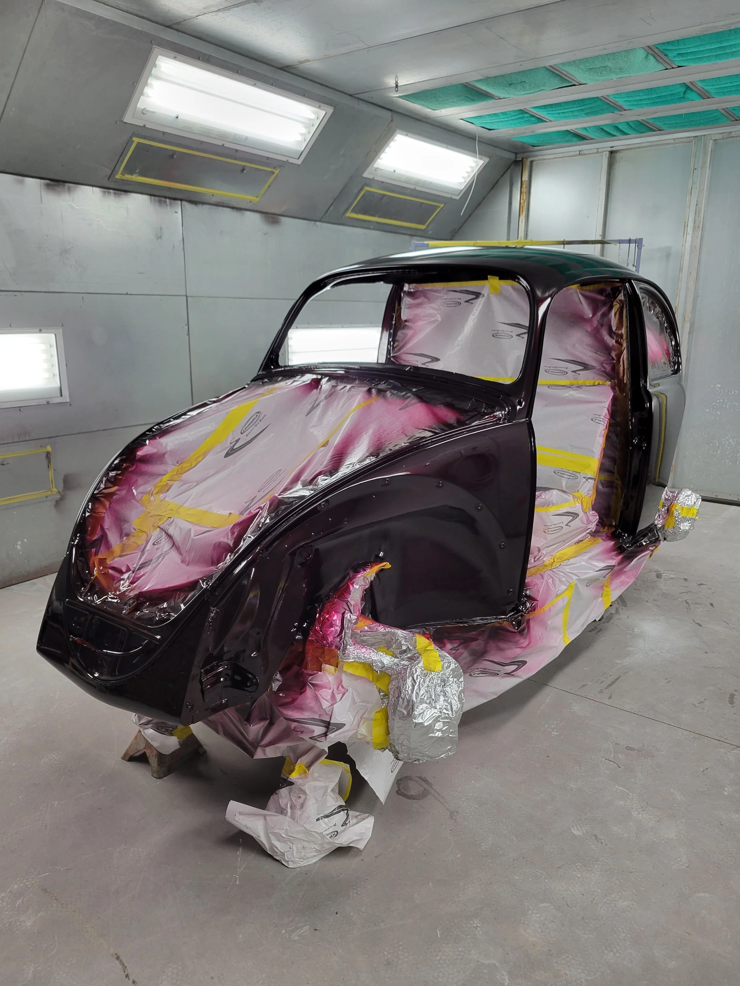 A black car during the painting process in a spray booth, covered with masking tape and paper to protect certain areas, with paint supplies and masking materials on the floor.