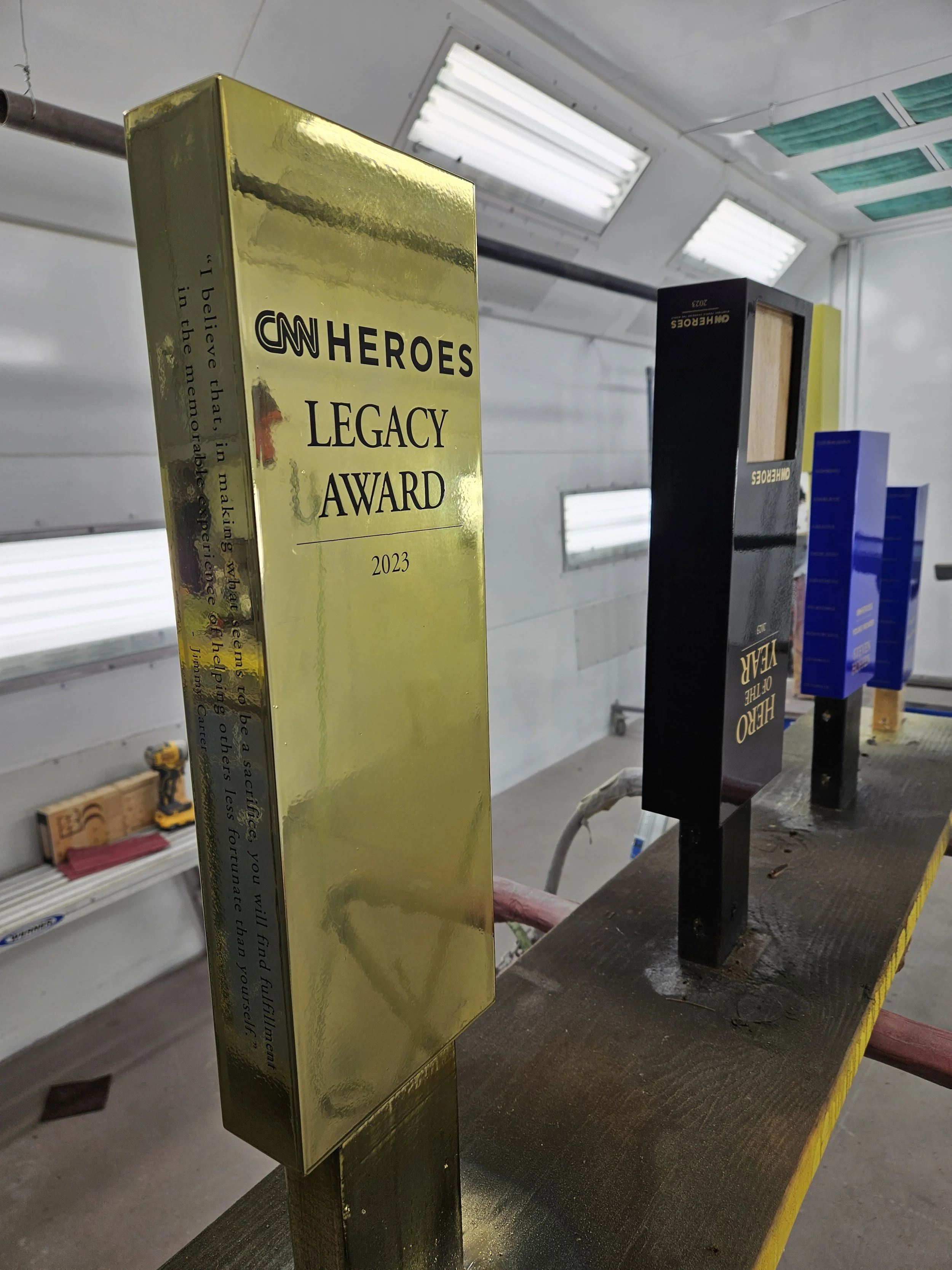 Golden and black CNN Heroes Legacy Award trophies on a wooden shelf in a workshop with fluorescent lights and tools in the background.