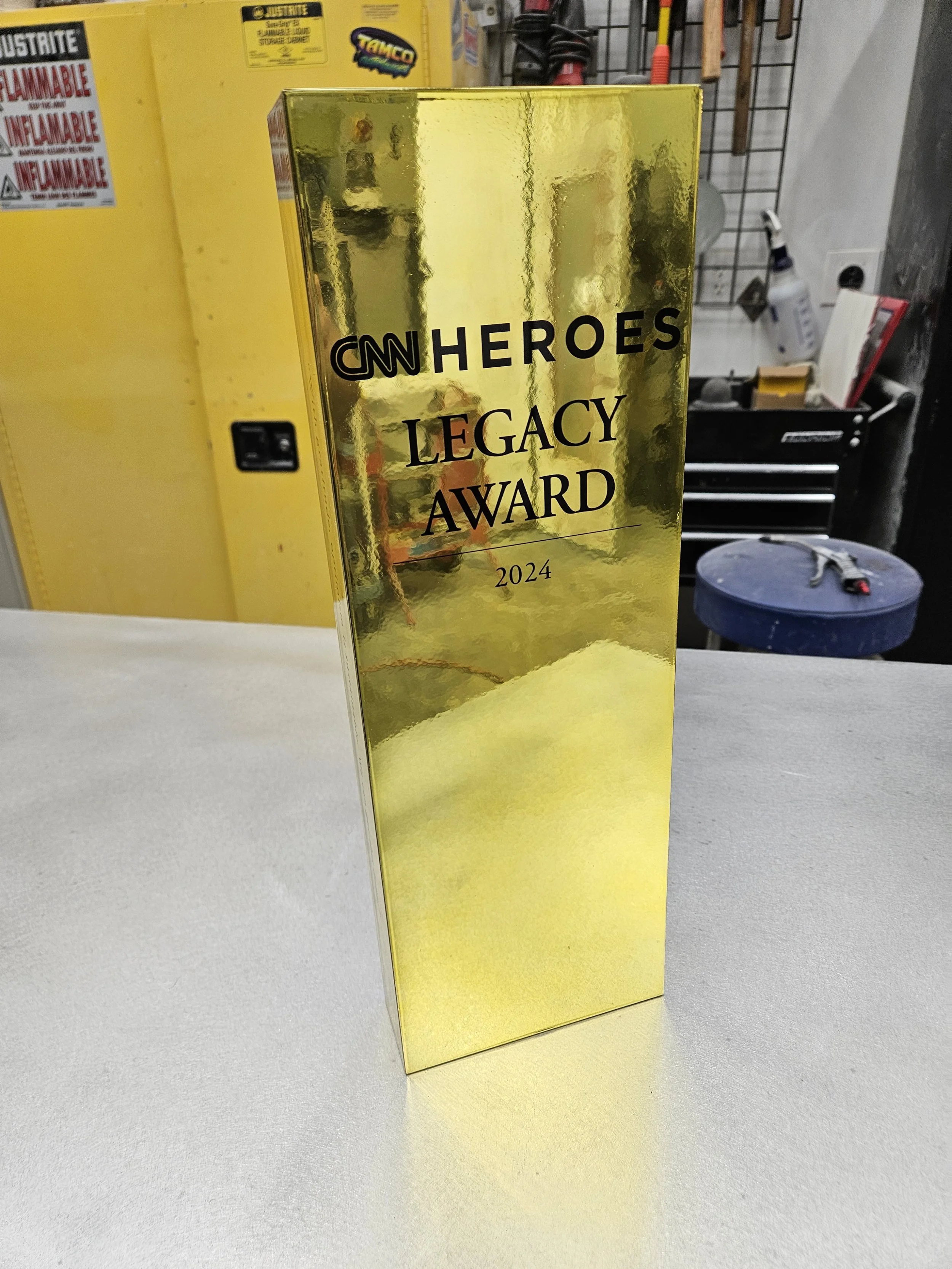 A shiny gold award trophy with black text reading 'CNN Heroes Legacy Award 2024' in a workshop or garage setting.
