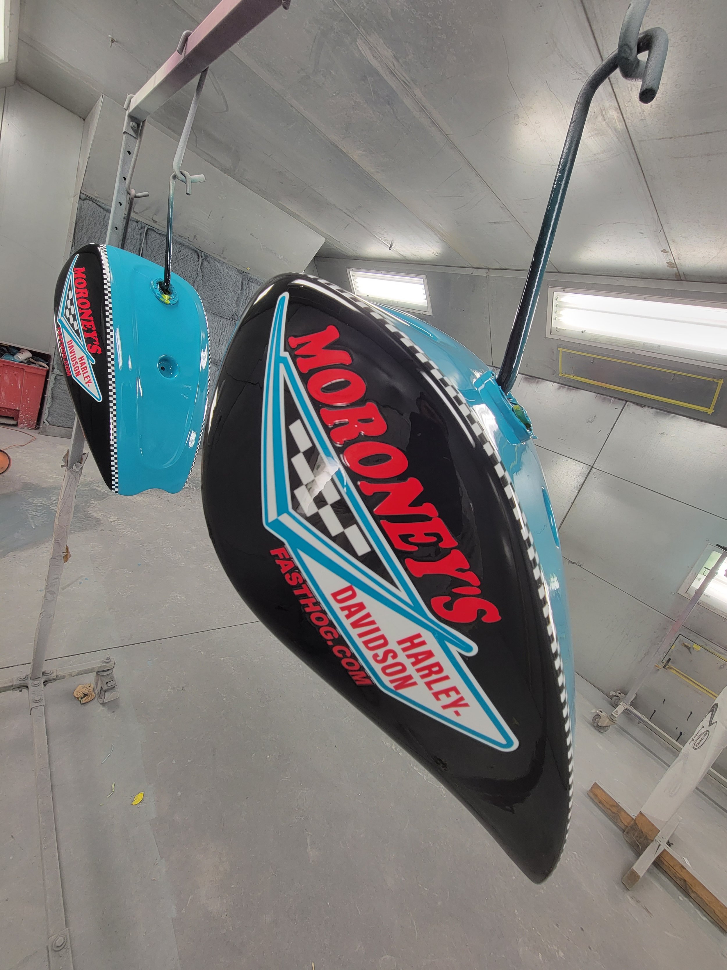 Decorated helmet with Harley Davidson and racing logos, hanging on a rack in a garage with fluorescent lights.