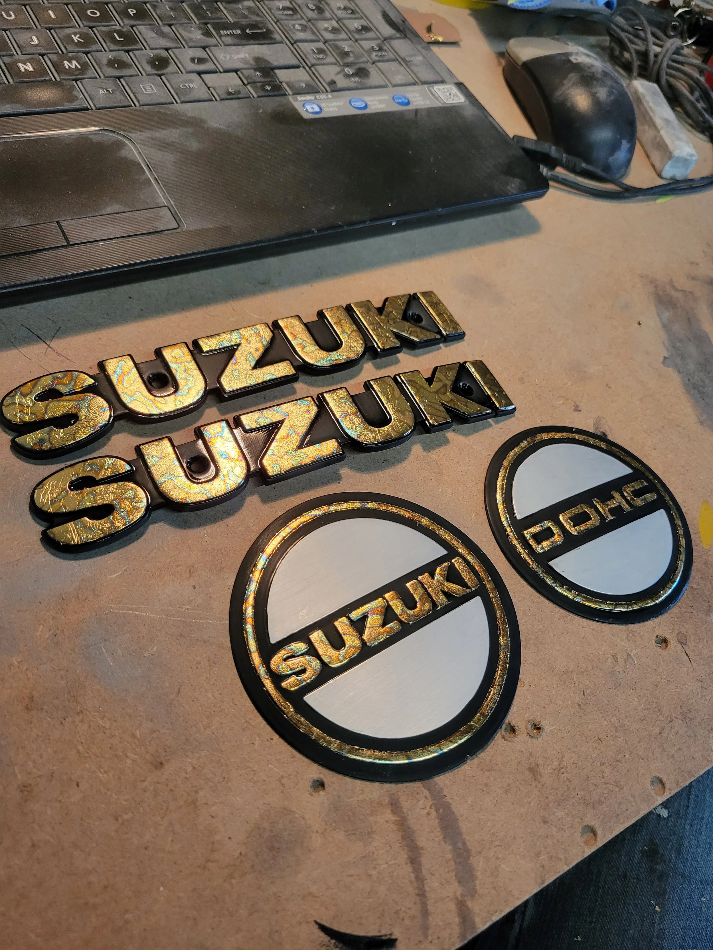 Two gold and black Suzuki badges and two Suzuki logos placed on a workbench, with a laptop, mouse, and scattered tools visible in the background.
