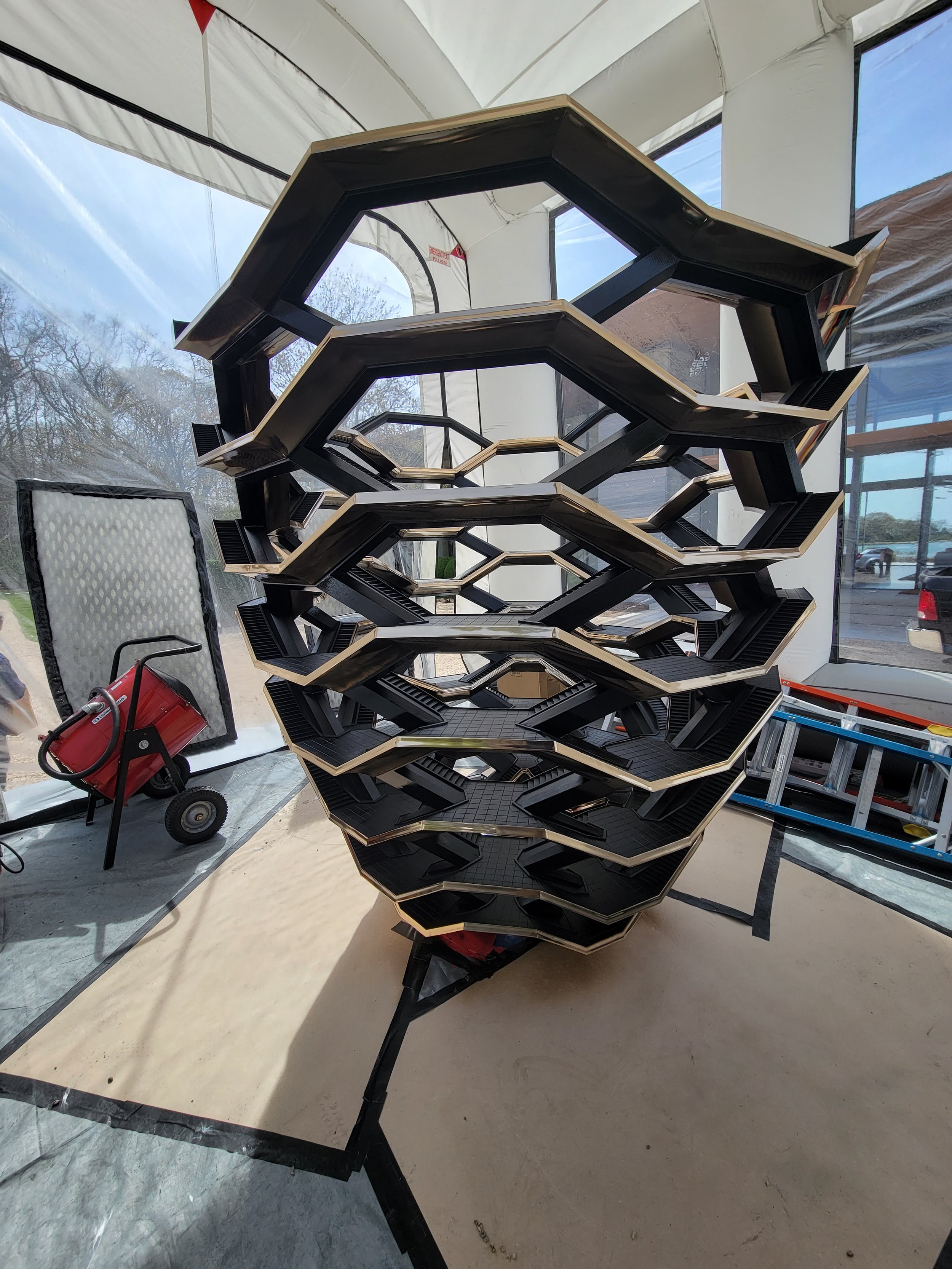 A unique black metal and beige shelving unit with multiple stacked, geometrically shaped tiers inside a bright, white-clad workspace with large windows.