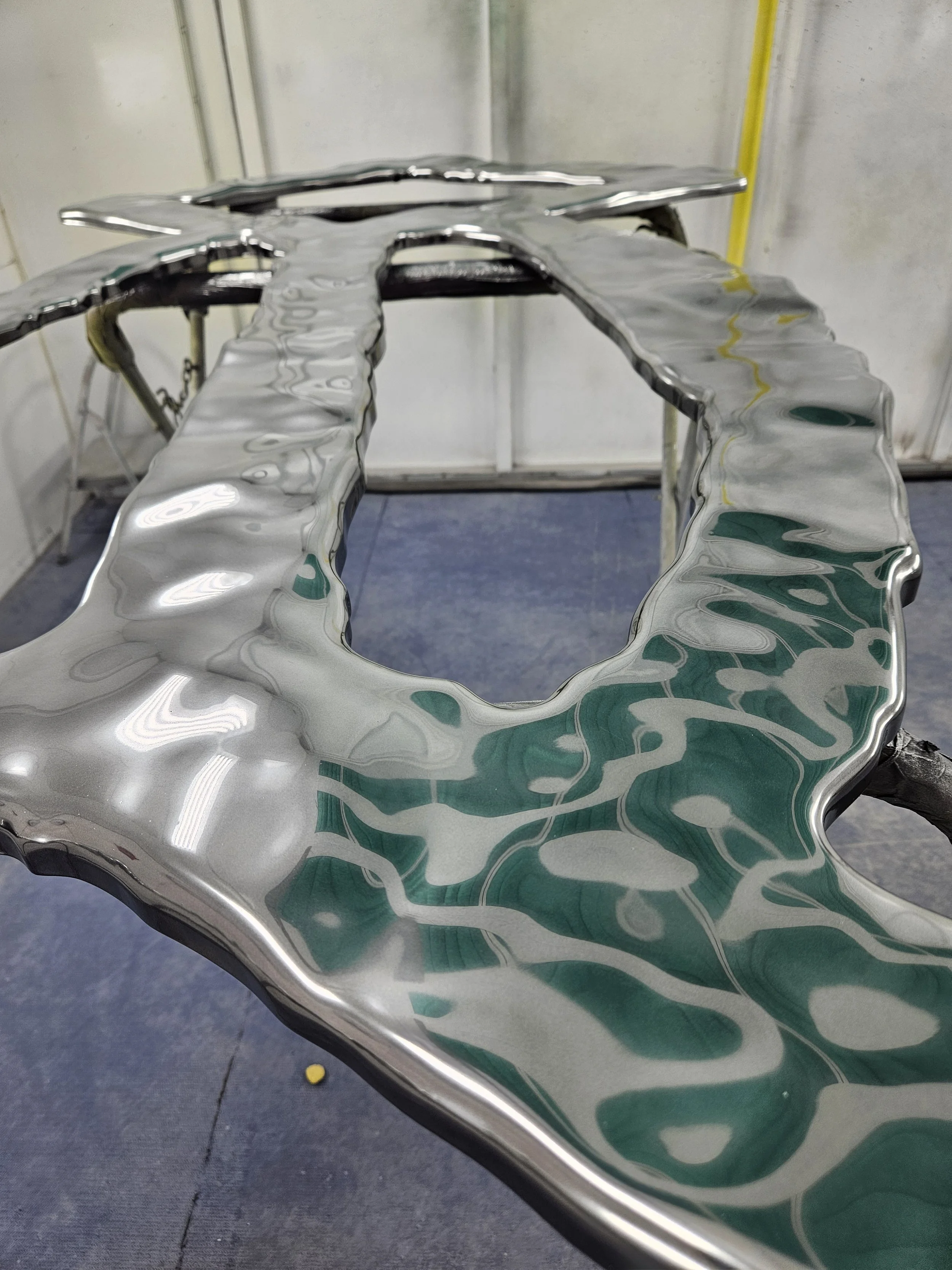Close-up of a shiny, metallic, reflective sculpture with a wavy, irregular surface, situated in an industrial or workshop setting.