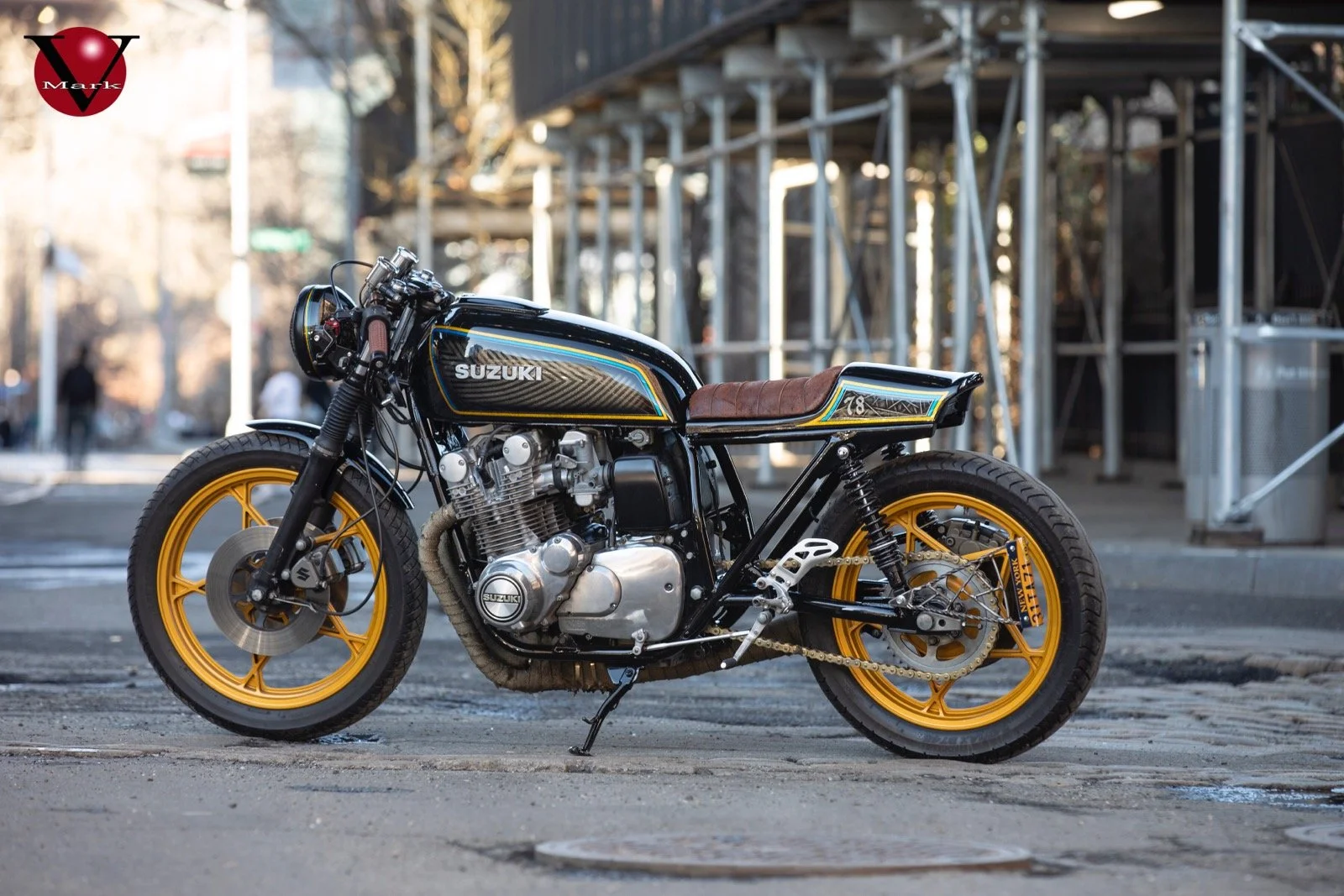 A custom Suzuki cafe racer motorcycle with black and yellow accents, parked on a city street near scaffolding and buildings.