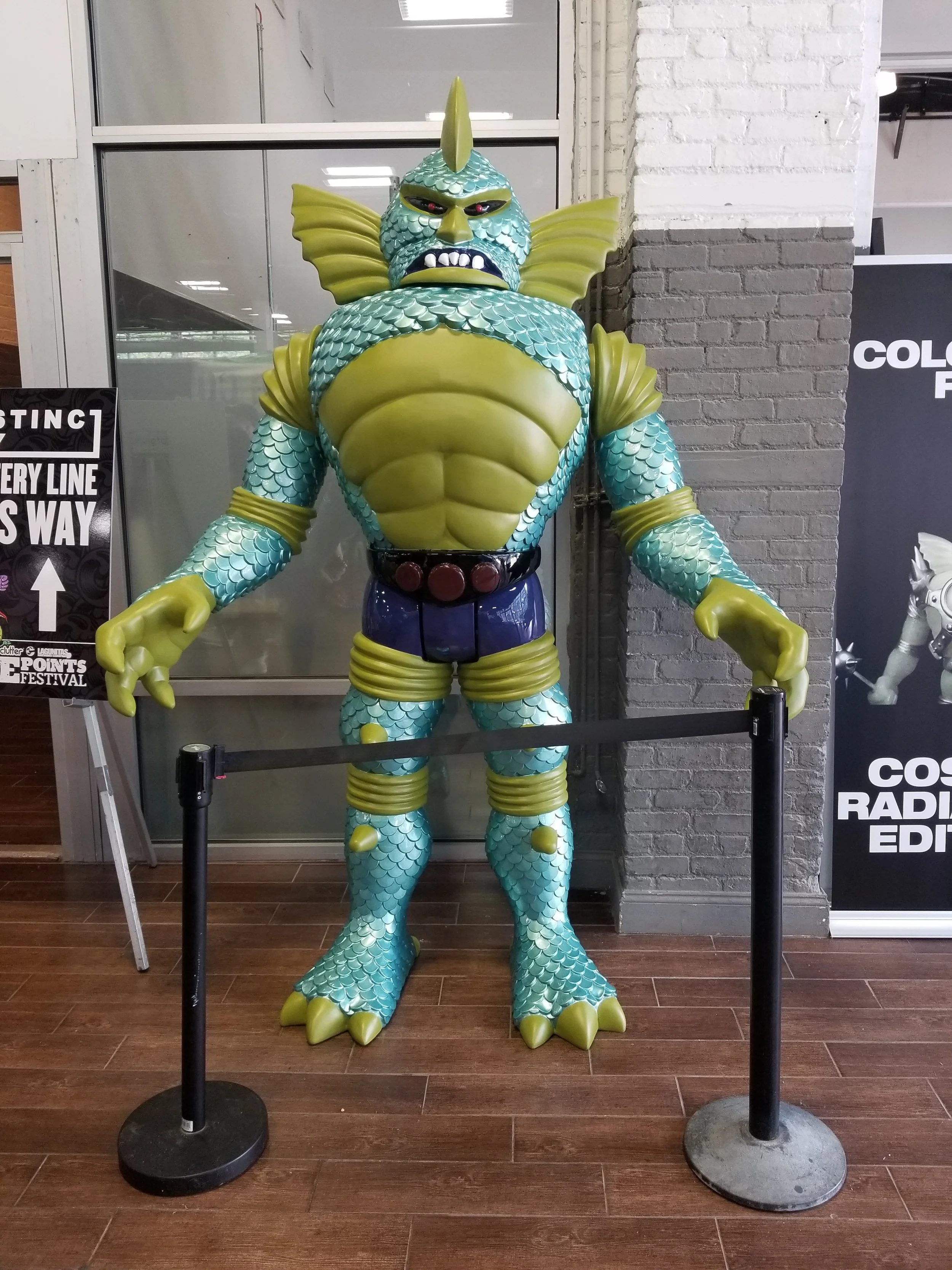 A large colorful statue of a mythical sea creature, resembling a fish-man hybrid, with green scales, large fins, and muscular build, standing inside a roped-off area in a store or exhibit.