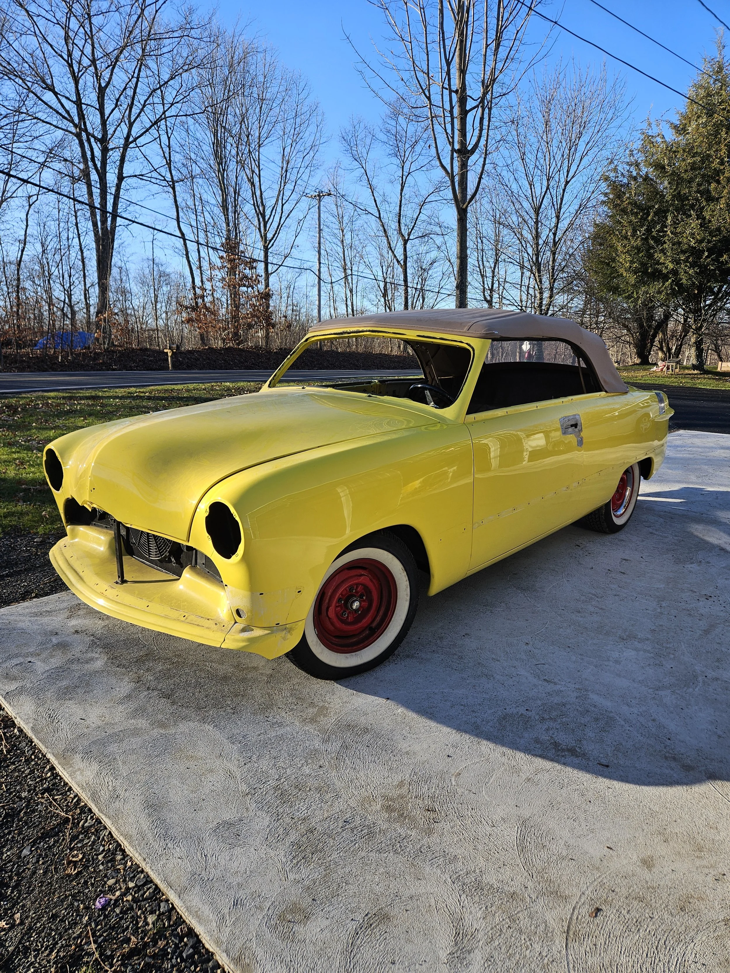 A vintage yellow convertible car with red wheels parked on a concrete driveway, missing headlights and front grille, with a background of leafless trees and a clear blue sky.