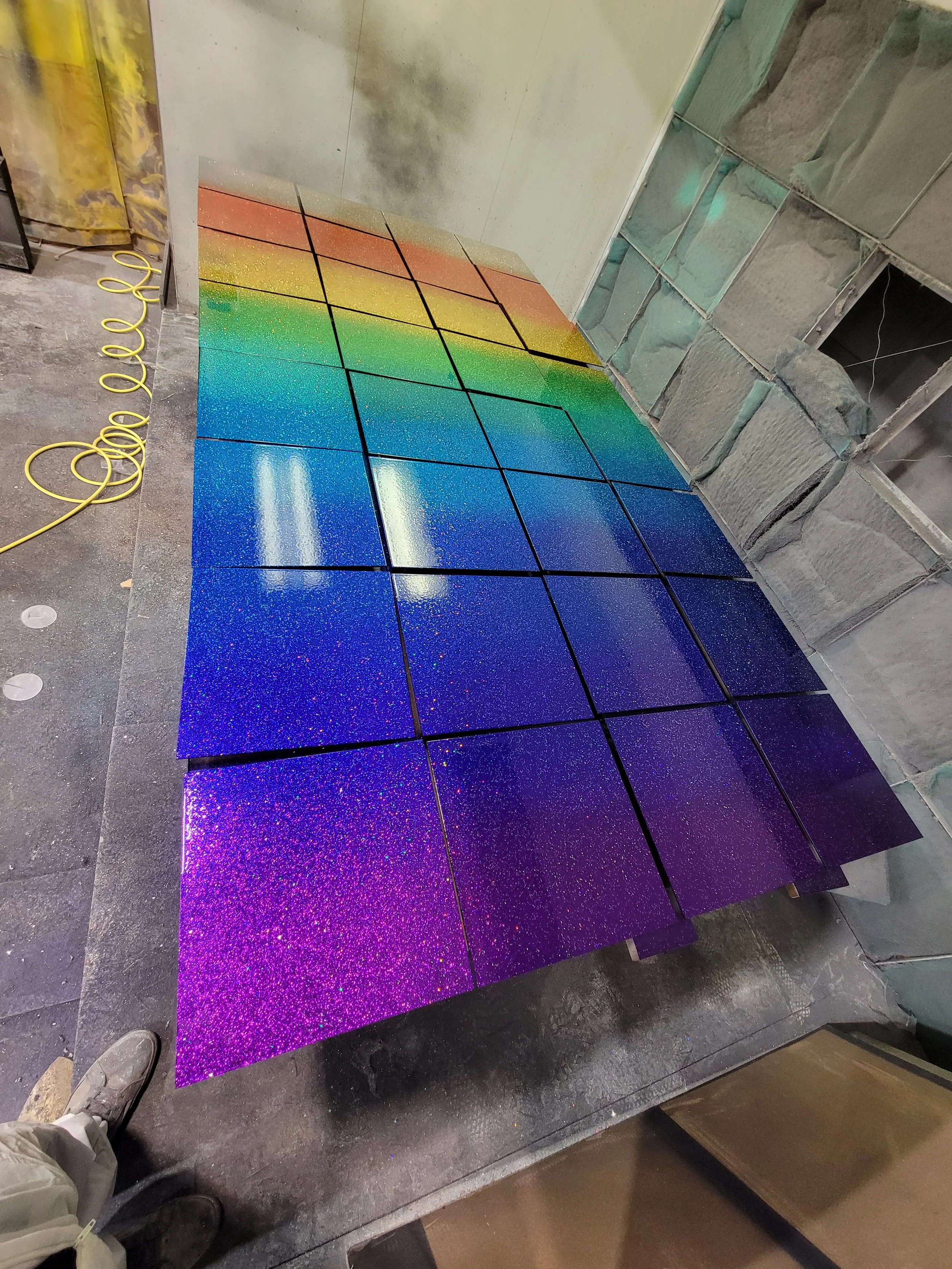 A floor with tiles painted in a gradient rainbow pattern, transitioning from purple to blue, green, yellow, orange, and red. The tiles are shiny with a speckled glitter finish.