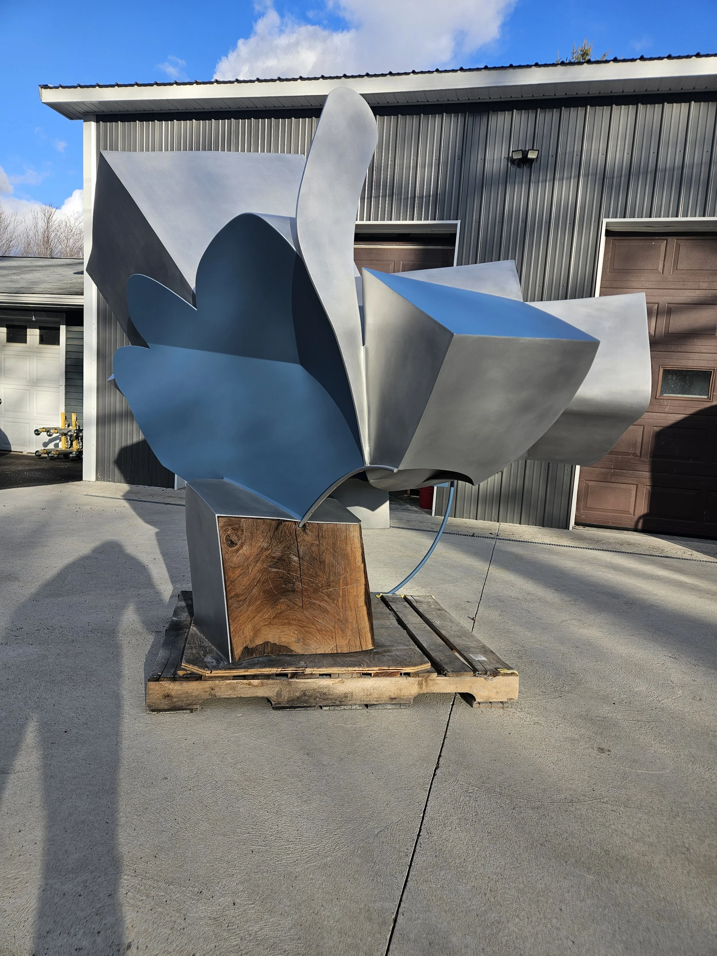 A modern abstract sculpture made of metal with geometric and organic shapes, mounted on a wooden base, outdoors on a concrete surface with a building and garage doors in the background.