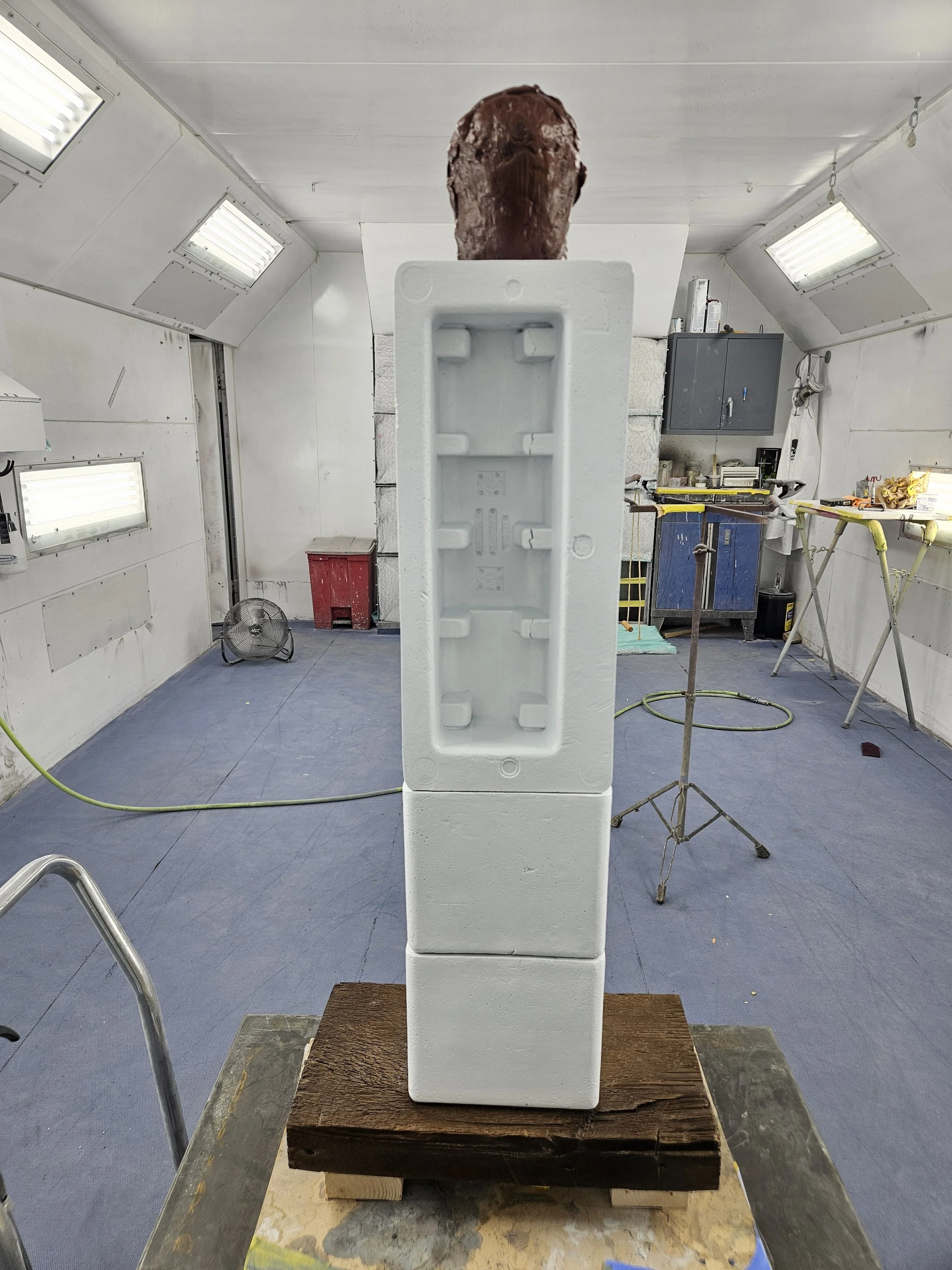 A foam sculpture of a human head on top of a rectangular foam base, placed in a workshop with various tools and equipment around.