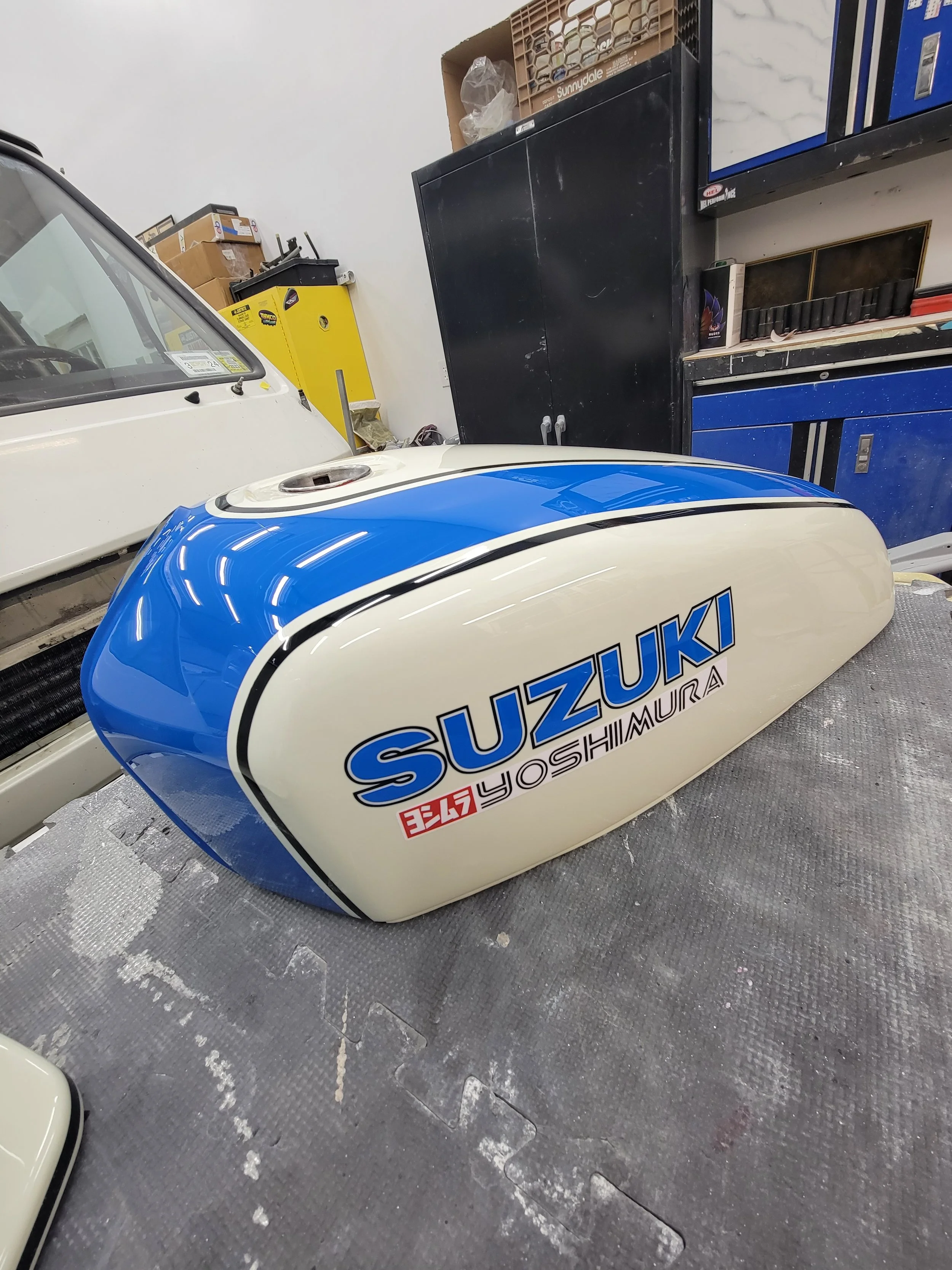 A vintage Suzuki motorcycle gas tank painted in cream and blue with Suzuki logo and Yoshimura branding, placed on a workbench in a garage.