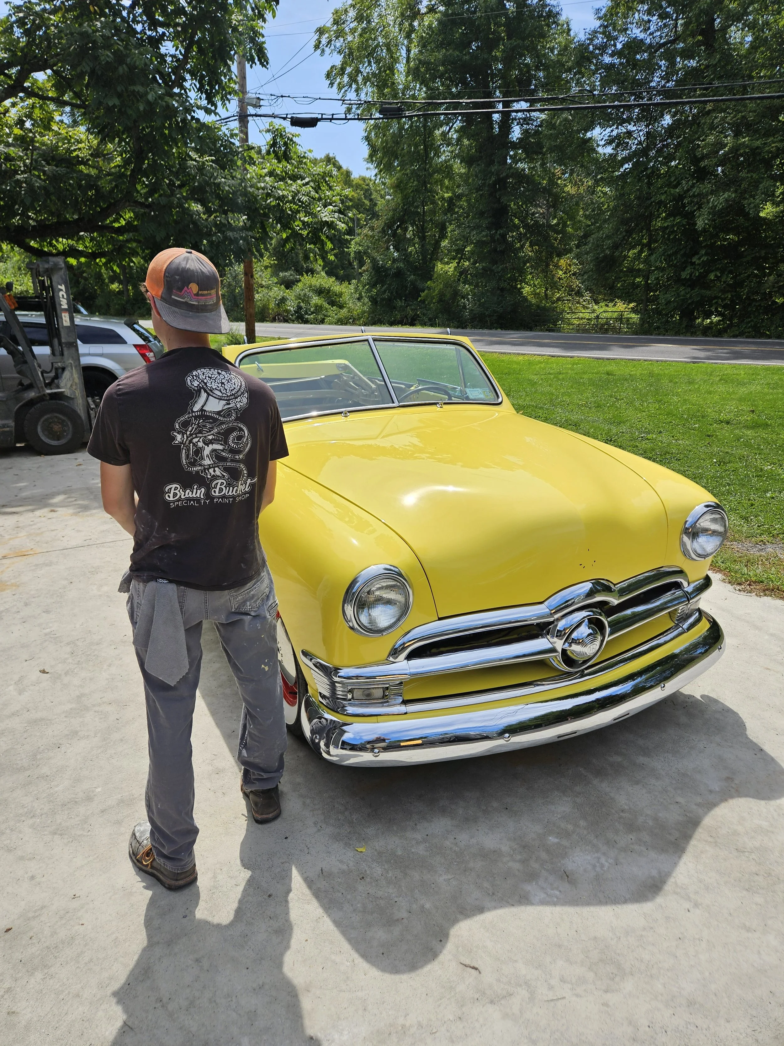 Person in a black t-shirt and gray pants standing next to a yellow vintage car on a concrete driveway, with green trees and a road in the background.